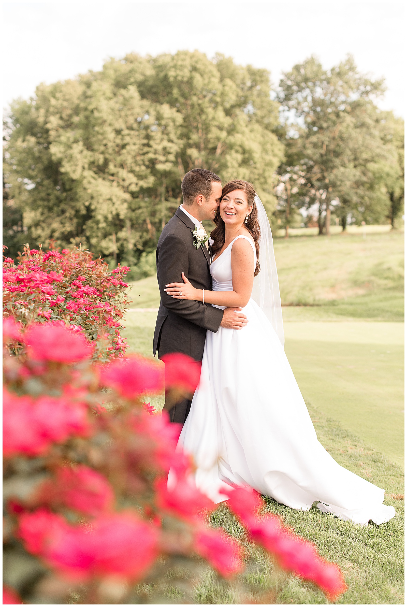 bride looking off to side and giggling as groom nuzzles in, in front of a rose bush