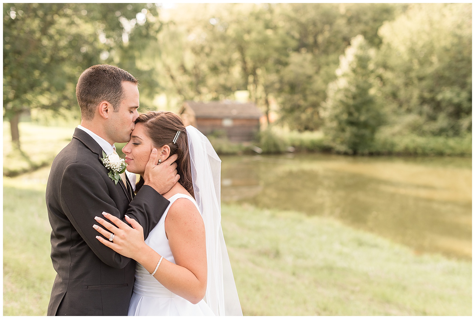 groom kissing brides forehead in front of pond with light trickling through trees behind