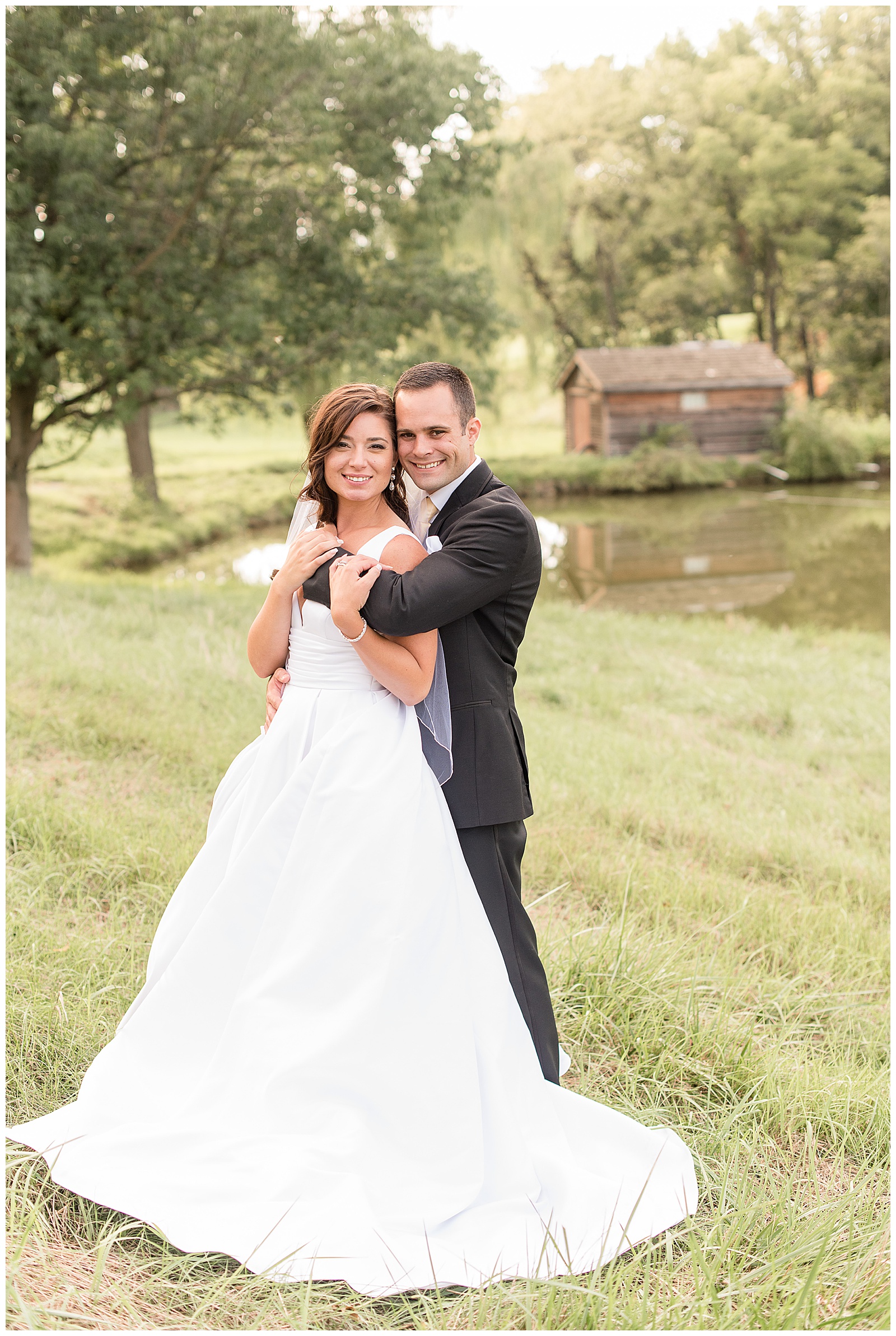 bride and groom in field in front of beautiful lake with groom having arms wrapped around bride
