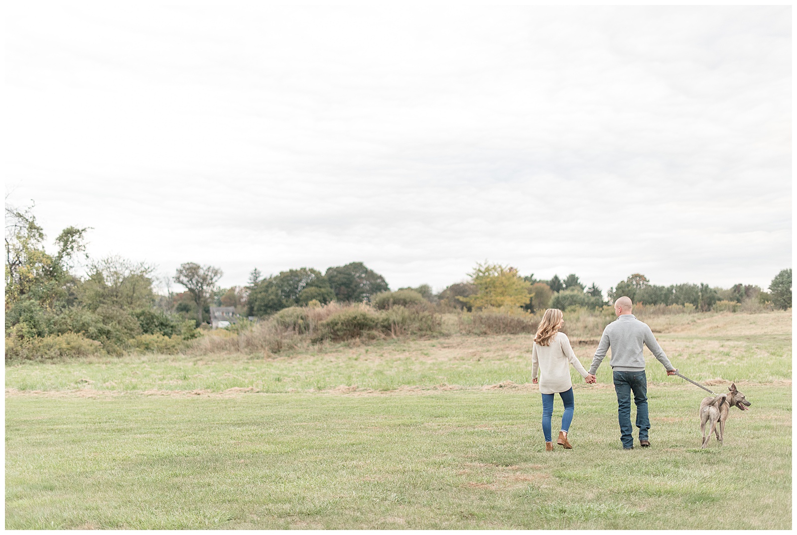 couple walking away from camera in open field with dog at side