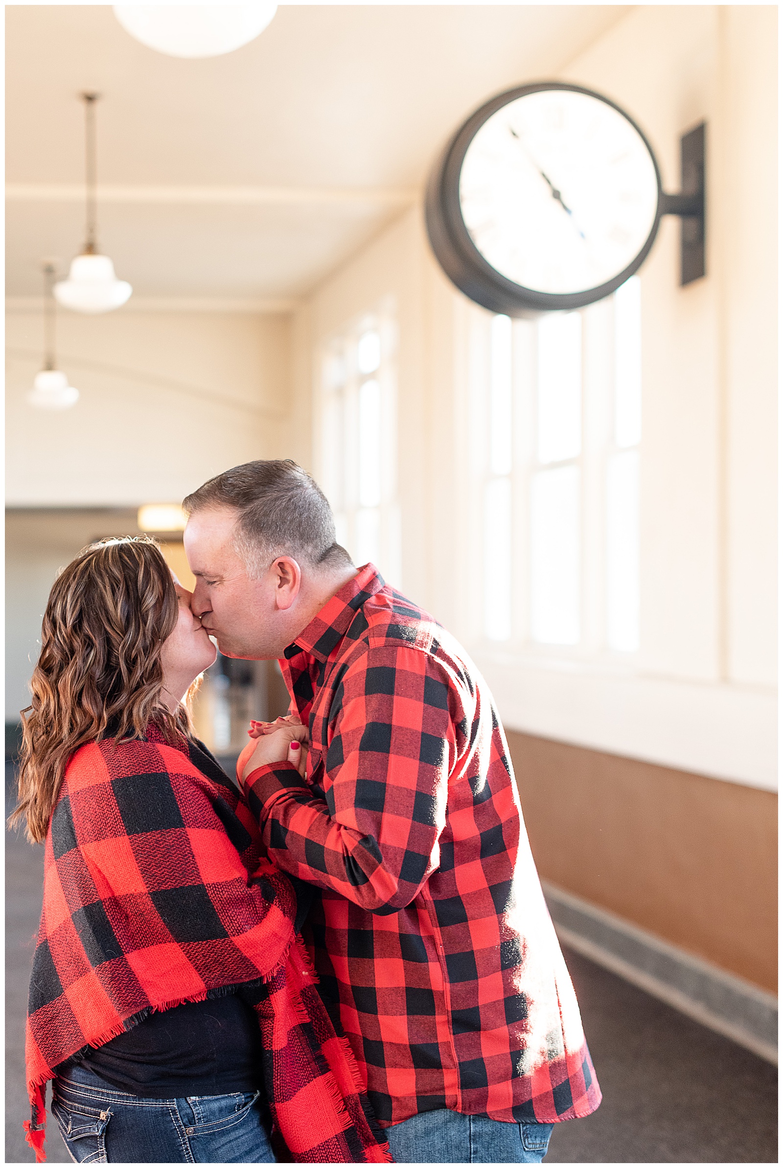 couple kissing inside train station with large clock hanging behind them