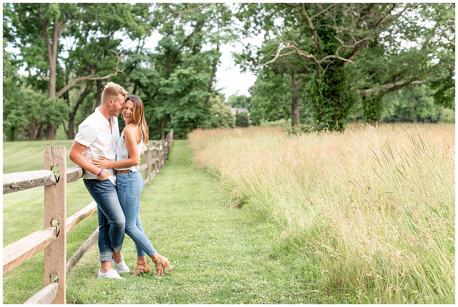 couple leaning against wood fence with tall grasses beside and girl smiles off camera while guy nuzzles in