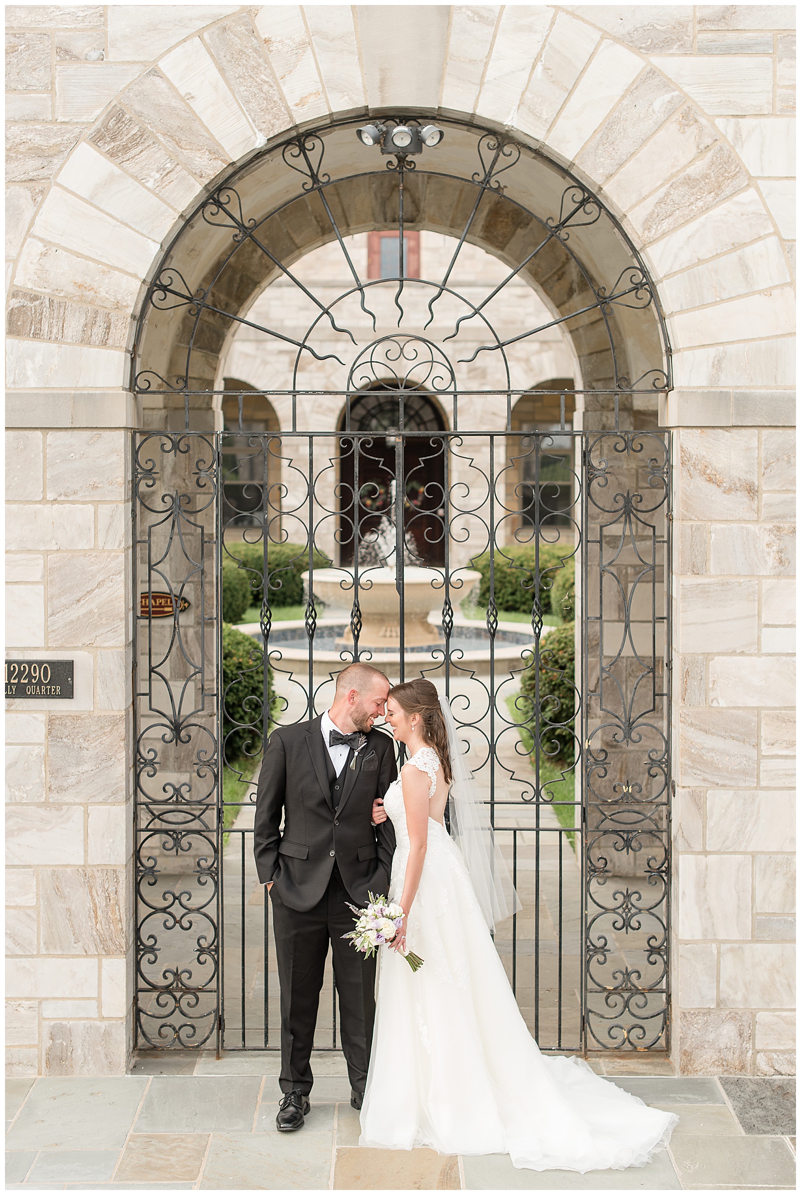 couple resting heads together standing in front of beautiful stone archway with iron gate