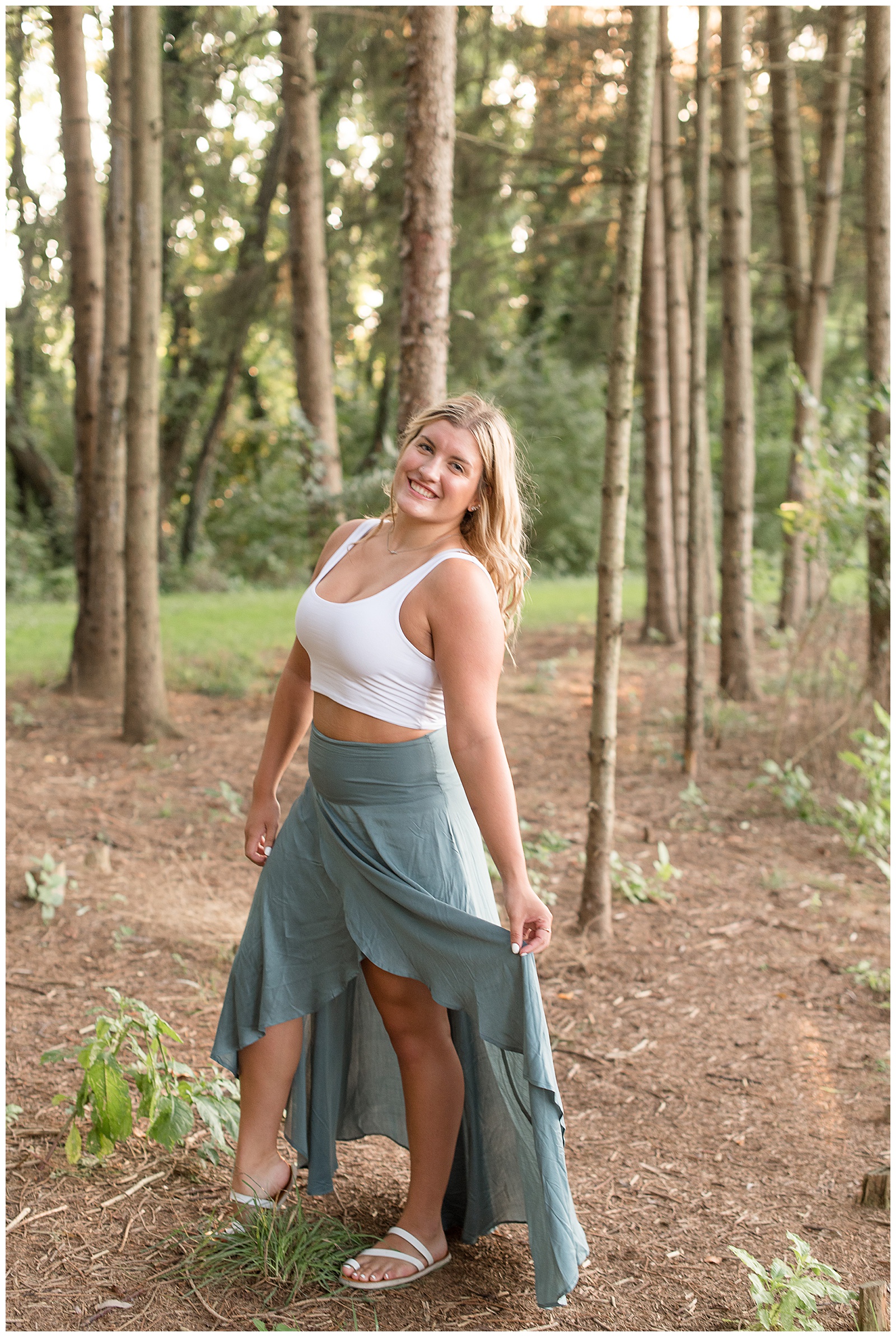 senior standing in middle of pine tree grove as she is holding her skirt and smiling at camera