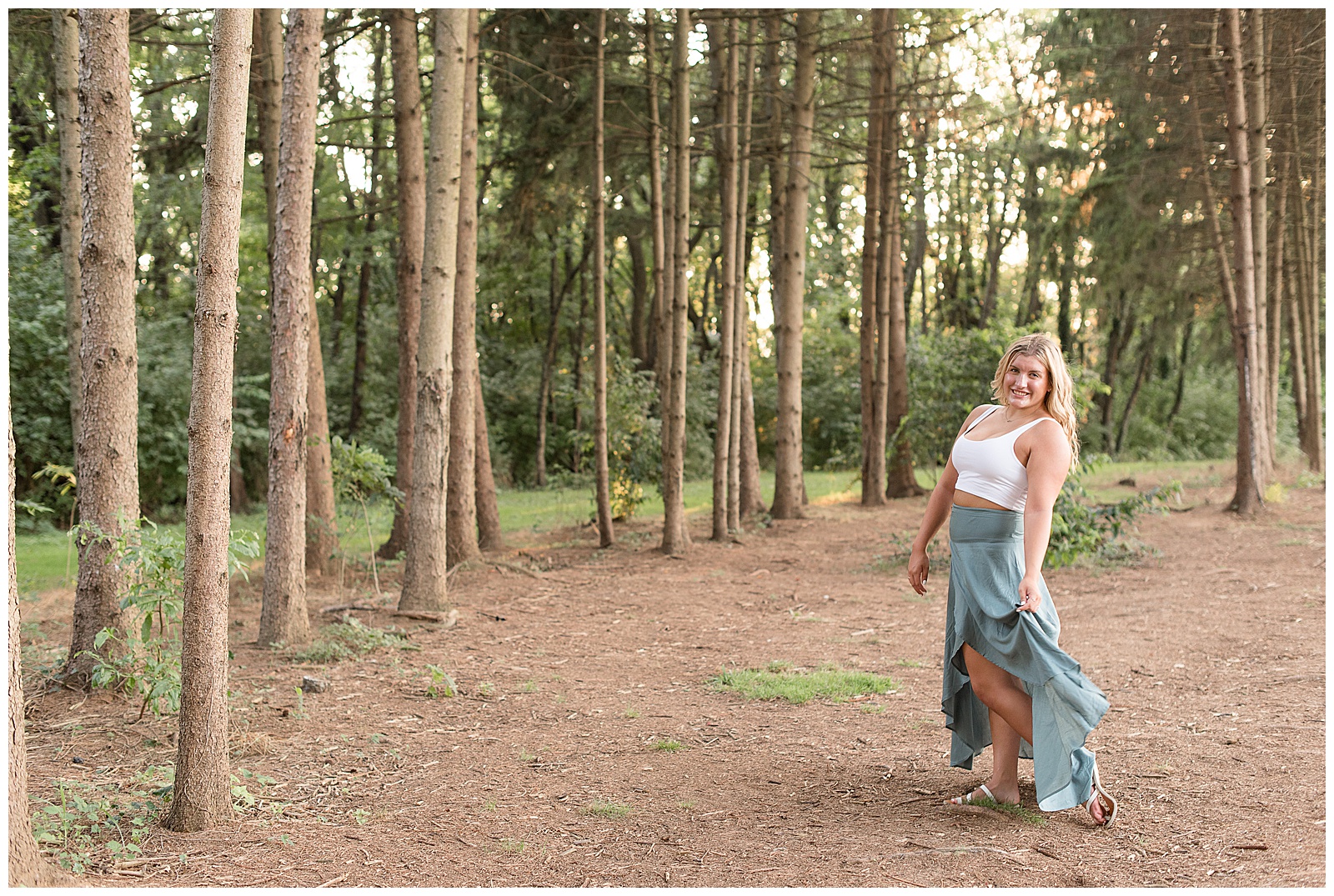 senior fluffing skirt in wind as she walks through grove of pine trees
