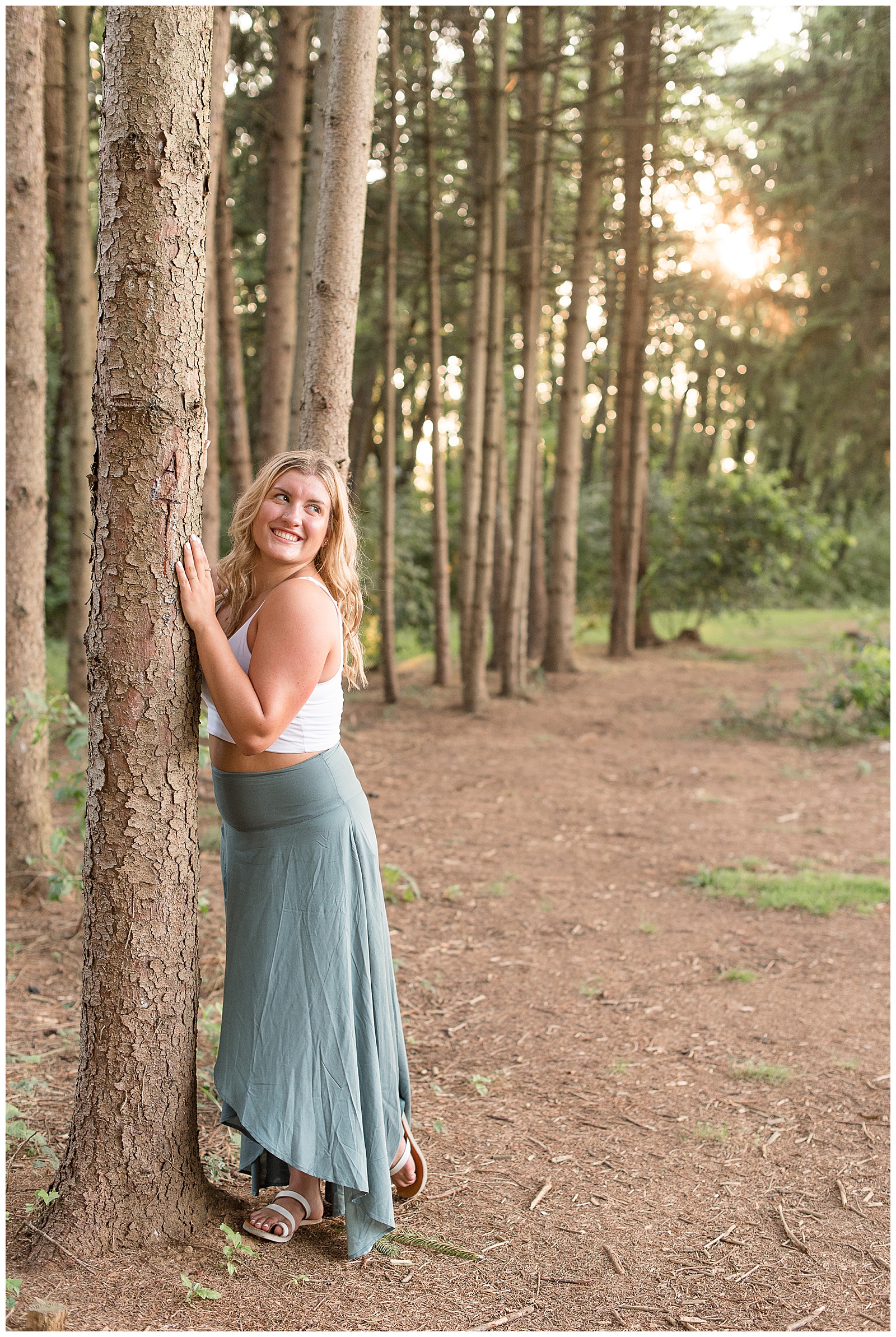 senior leaning on tree smiling over her shoulder with light coming through pine trees around her