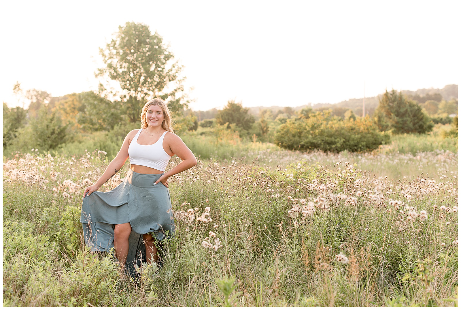 senior standing with hand on hip and other holding her skirt out surrounded by field of tall grasses