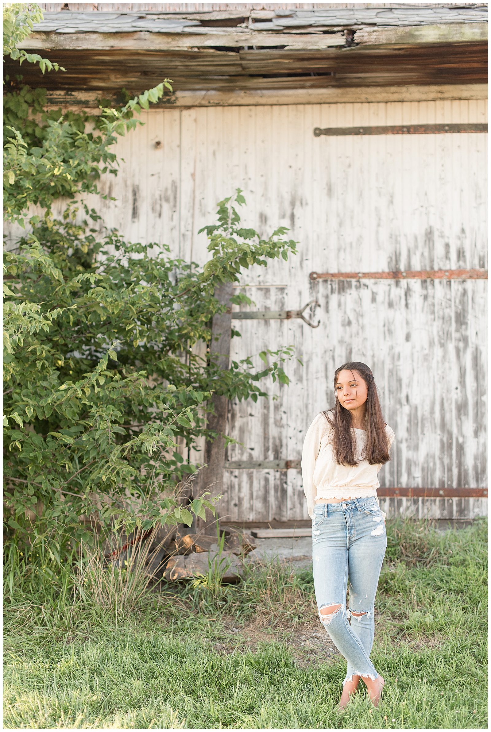 senior looking softly at camera holding hands together behind her back with feet crossed at ankles in front of barn door
