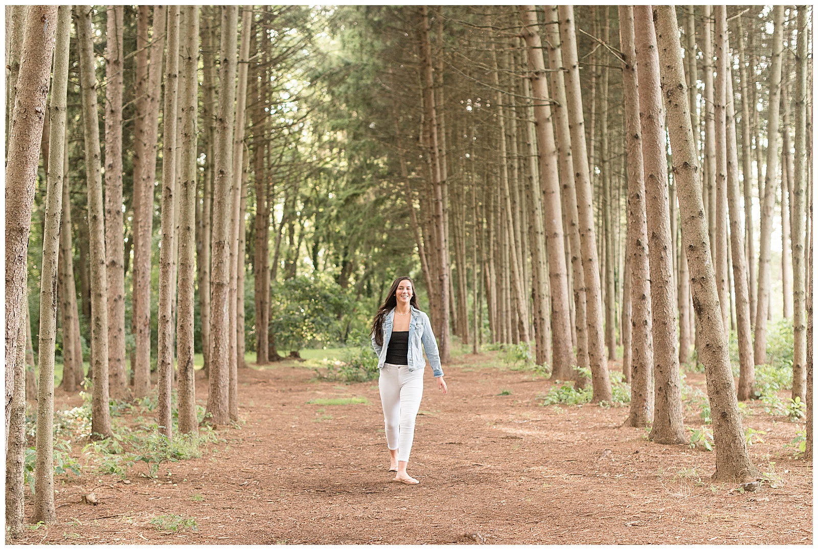 senior in white pants walking between rows of grove trees smiling at camera