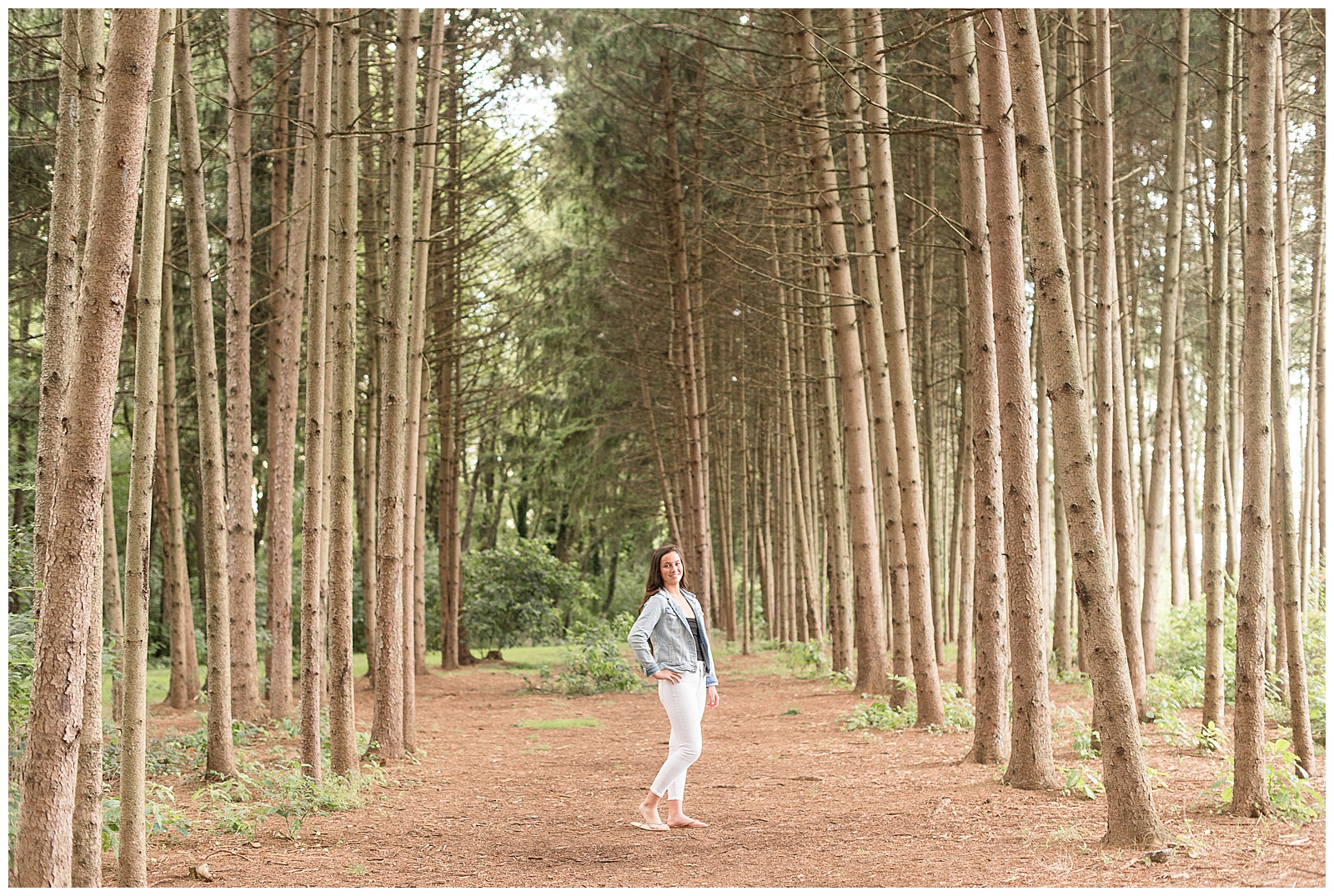 senior with hand on hip looking over shoulder smiling at camera with grove of pine trees around