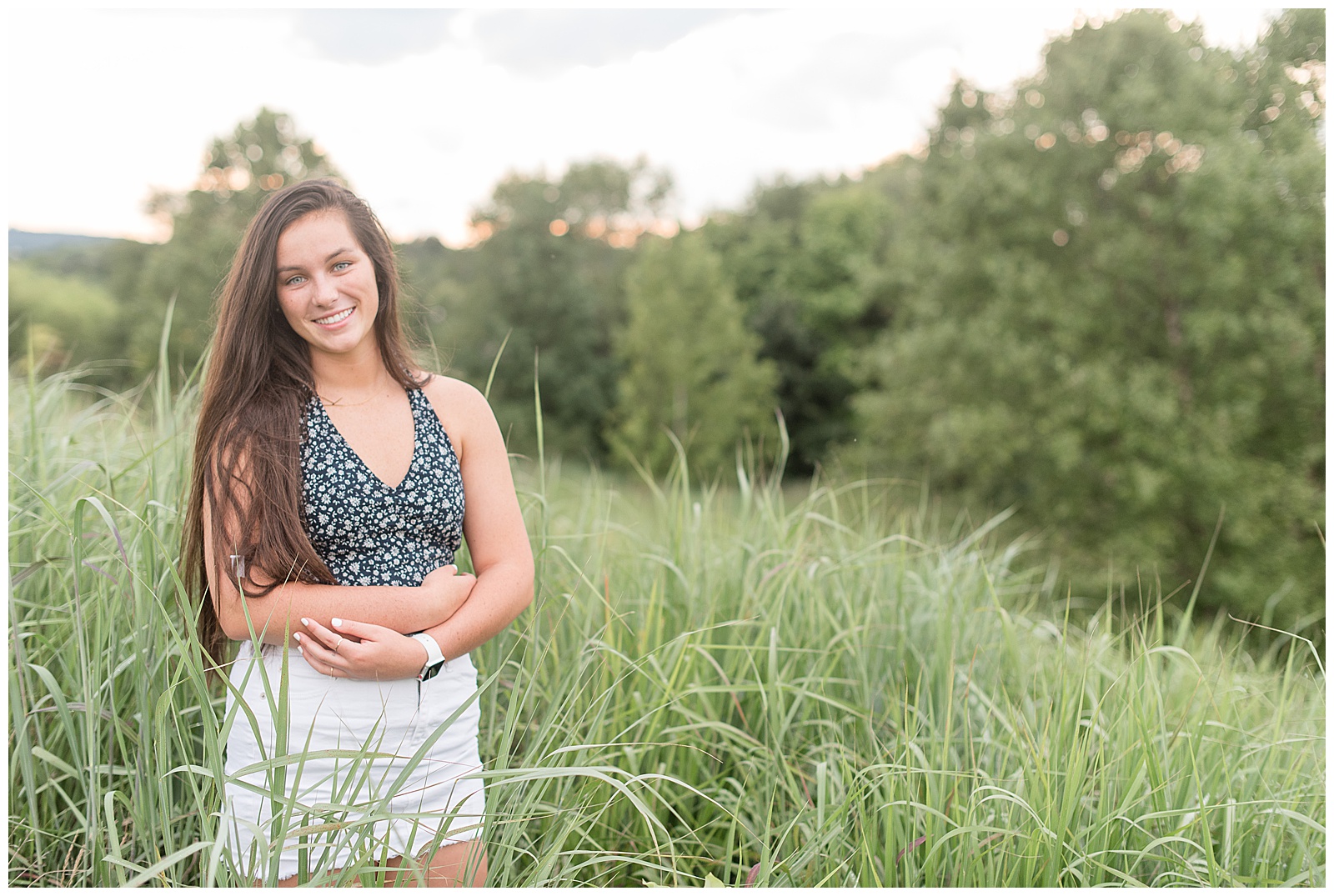 senior standing among tall grasses with white skirt and floral shirt smiling at camera