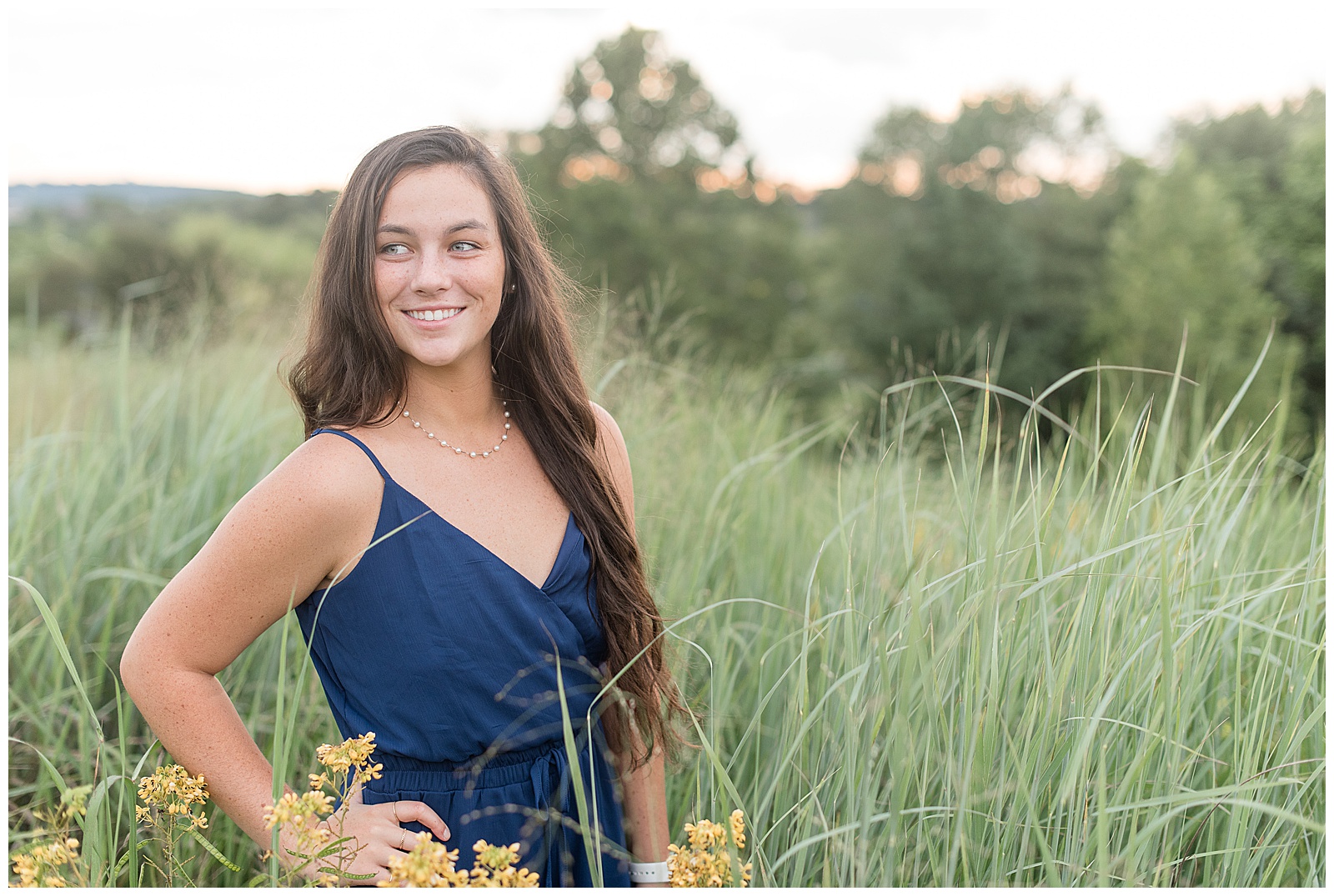 senior smiling off to side of camera with hand on hip in a blue romper with talll grasses around at Overlook Park