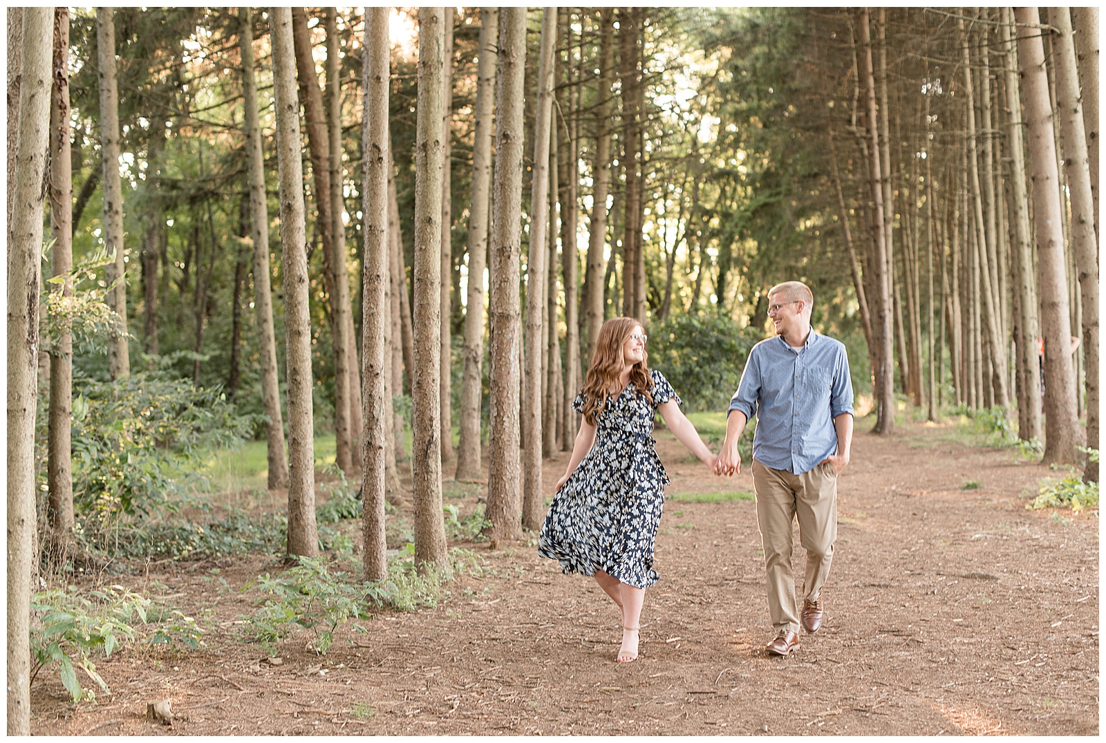 couple walking in a grove of pine trees smiling at each other while girl holds onto dress as it blows in the wind