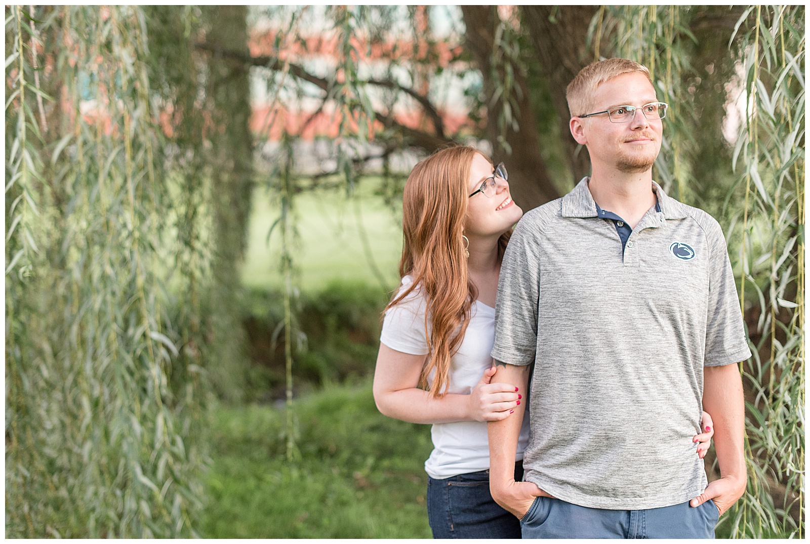 couple standing in front of willow tree with girl hugging guy from behind smiling at him as he smiles off to side