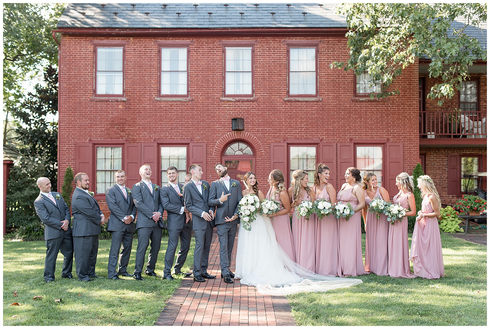 full bridal party smiling at each other and laughing standing in front of red brick house in courtyard