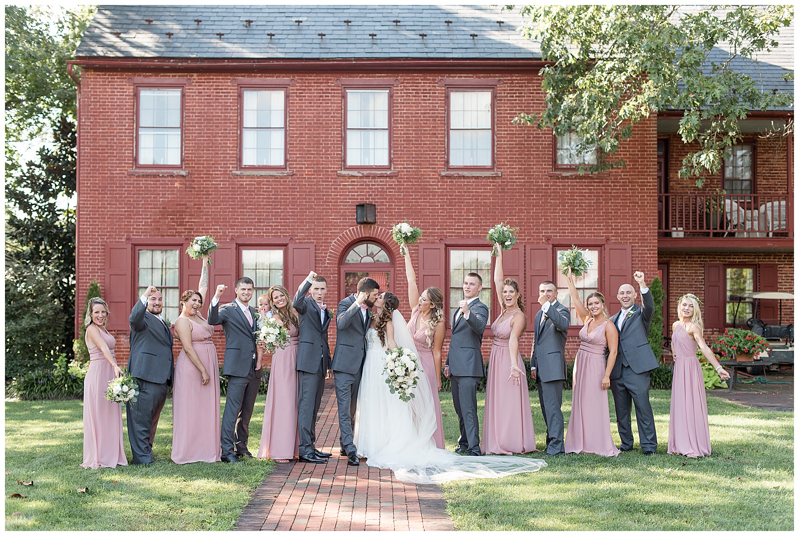 wedding party cheering as bride and groom share kiss in courtyard at Country Barn with red brick house behind