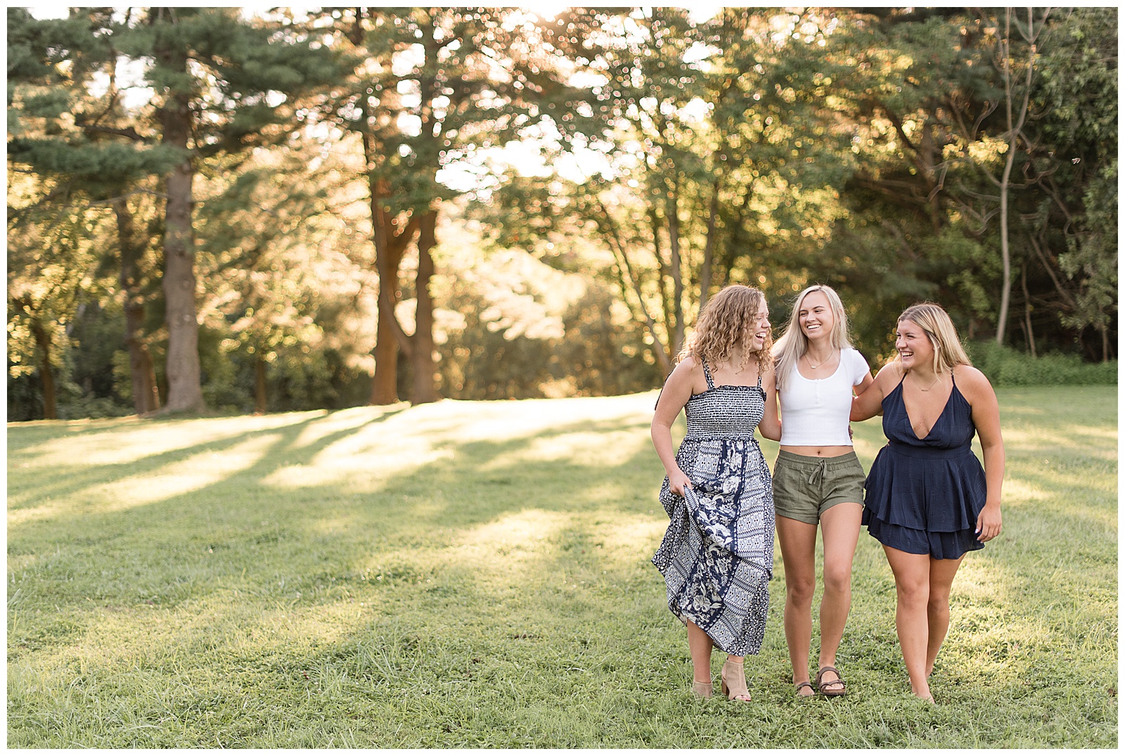 three senior girls walking forward together with arms around each others' backs while smiling towards each other with sunshine in the trees behind them in grassy field