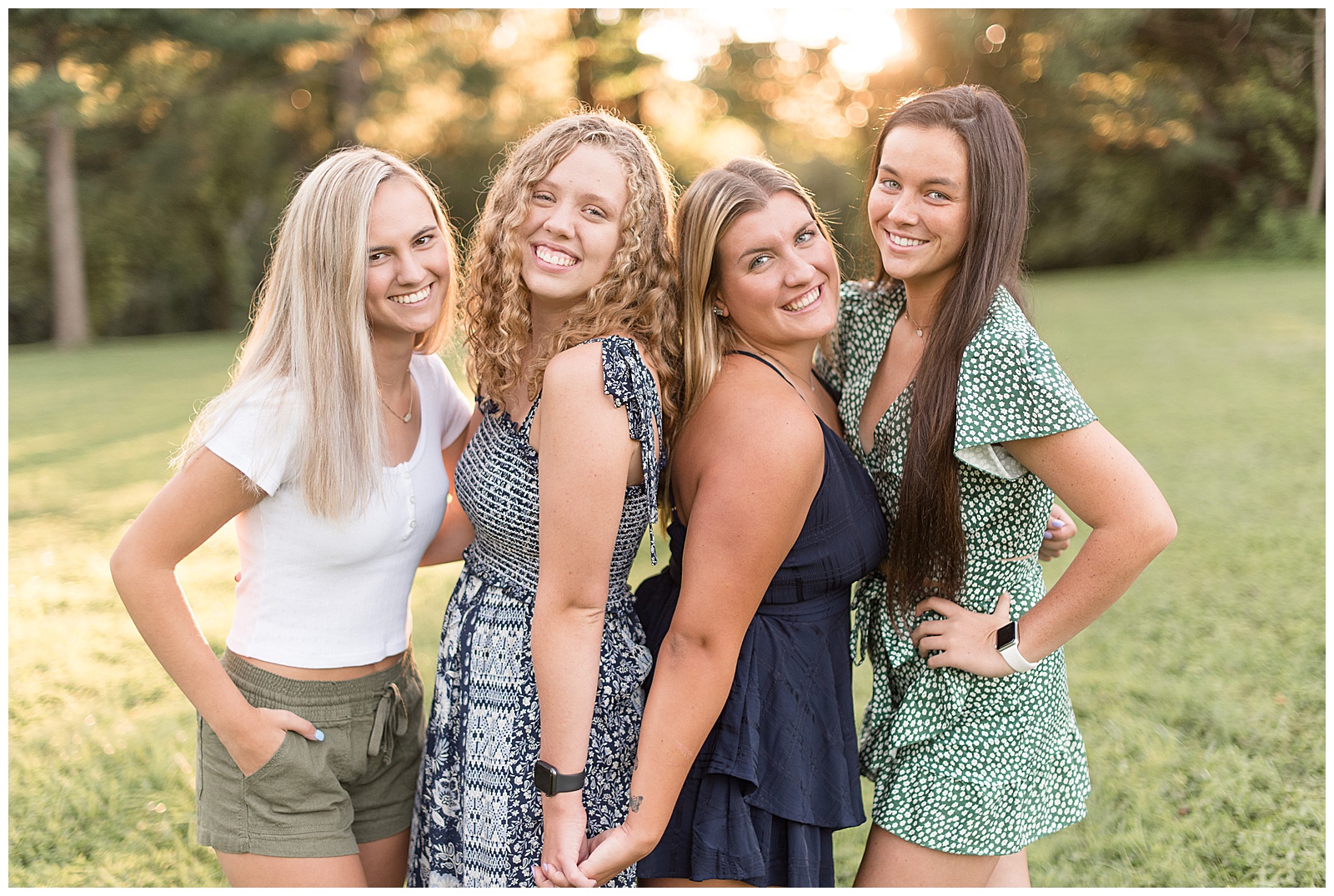 a close-up photo of all four senior girls facing one another smiling at the camera with the two girls in the middle having their backs together with their hands joined and the two girls on the ends having their hands on their hip or in their pocket with sunlit trees behind them