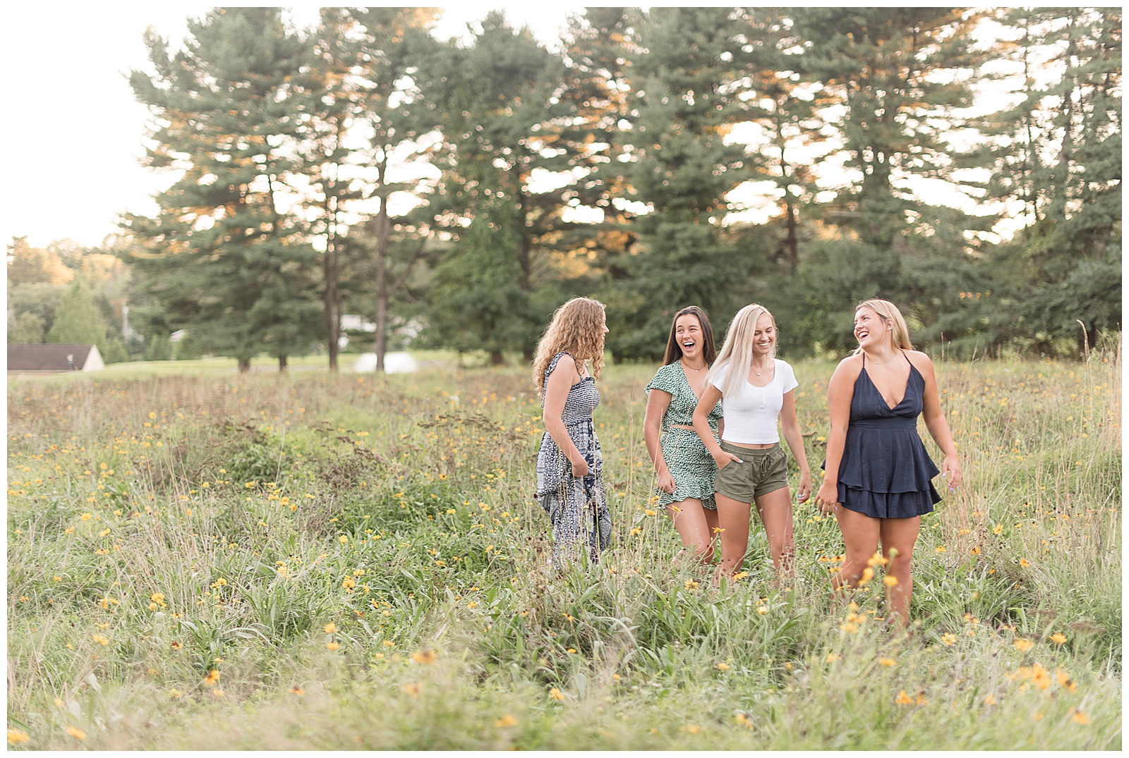 four senior girls walking together facing each other smiling and laughing together while coming towards the camera in the wildflower field with evergreen trees behind them