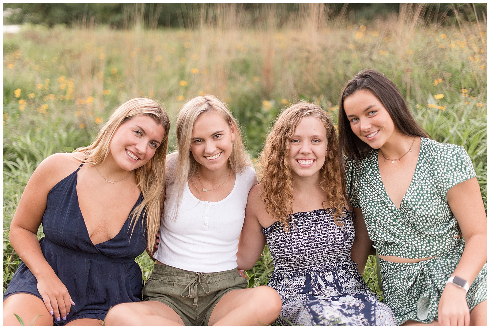 a close-up of all four girls sitting together in the wildflower field with their arms around each other and smiling at the camera