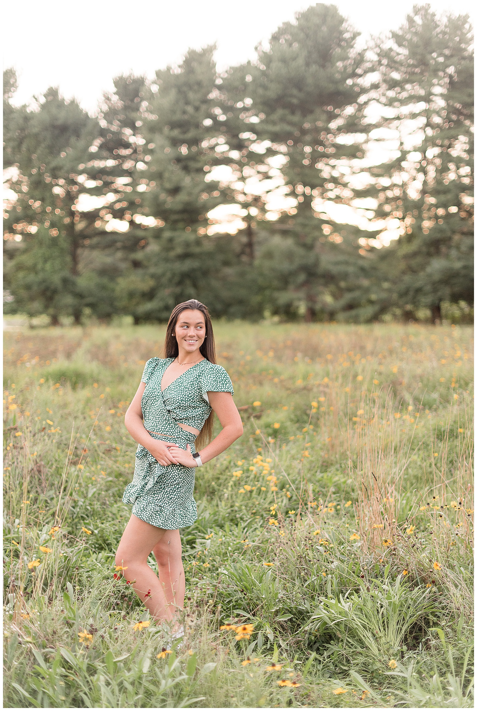 one senior girl in wildflower field facing right with both hands on her left hip while smiling over her left shoulder with evergreen trees behind her