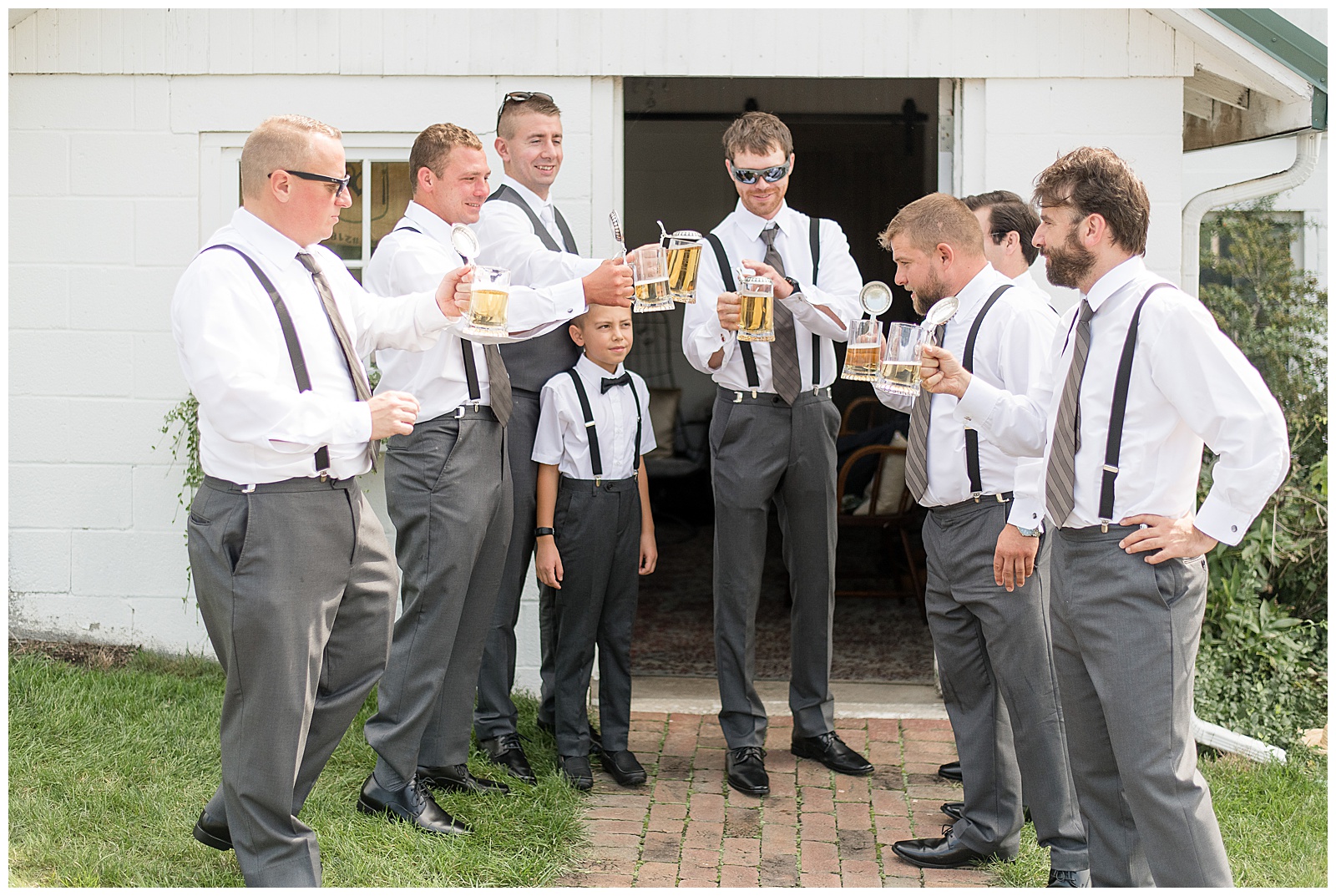 close up photo of the groom, groomsmen, and ring bearer standing in from of white barn on brick pathway toasting one another with drinks and everyone facing one another and smiling in their gray dress pants, white dress shirts, ties, and black suspenders at Lakefield Weddings in Manheim, PA