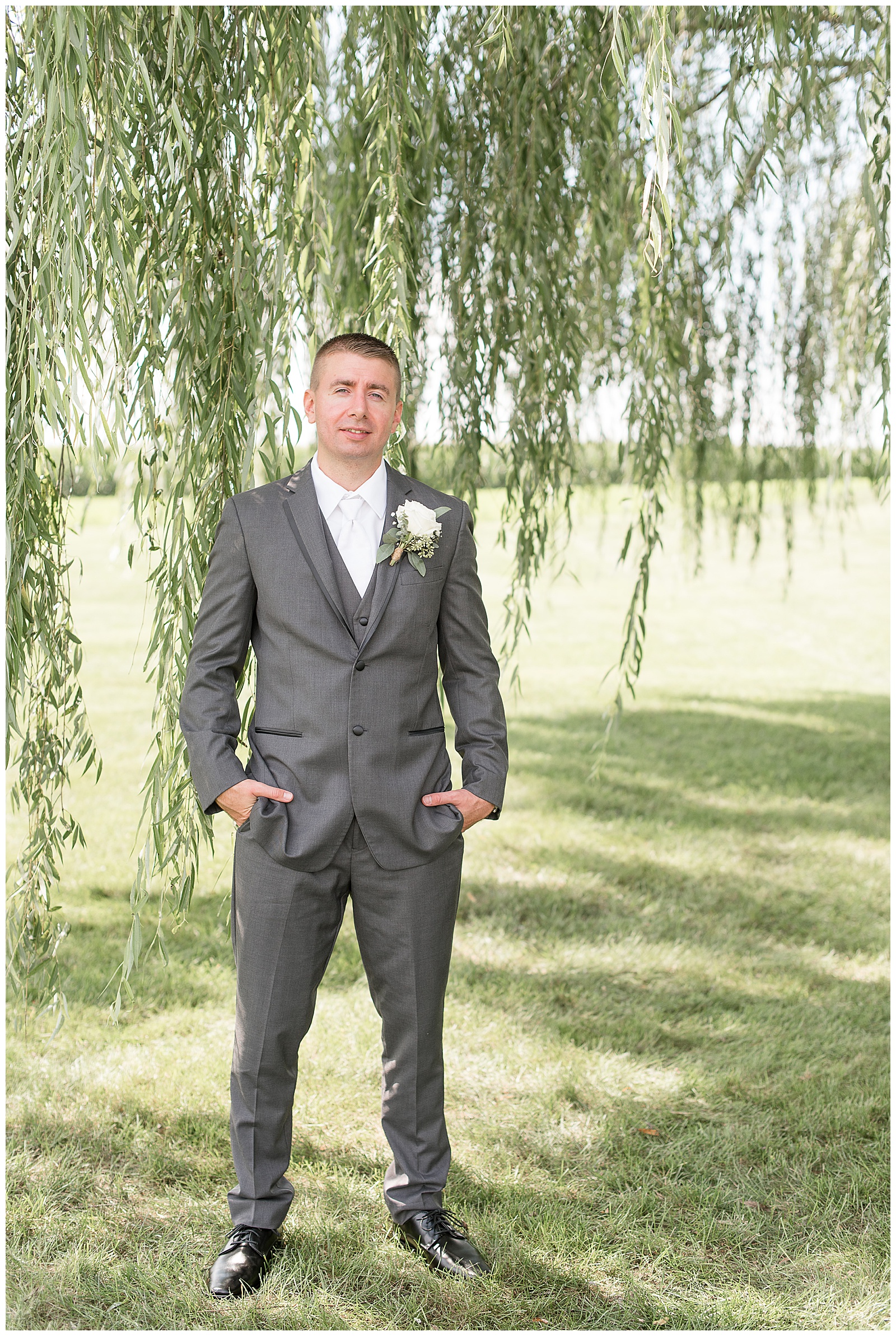 close up photo of the groom facing the camera in his dark gray suit with both of his hands in his pockets smiling at the camera in a large grass field in front of a willow tree on a sunny day at Lakefield Weddings in Manheim, PA