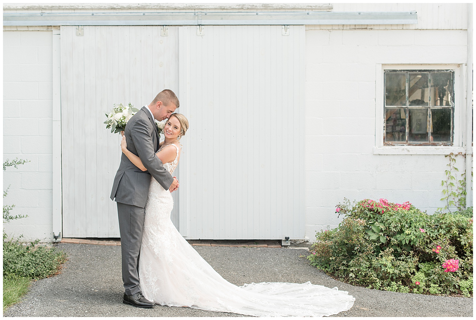 the bride and groom are facing each other with the groom's forehead resting on the right side of her head as she faces the camera and smiles and they're standing on the left side of the photo with the long train of her dress spread out across the driveway in front of the white barn with potted flowers on the right side of the photo on the driveway and the bride is holding her bouquet of flowers behind the groom's back at Lakefield Weddings in Manheim, PA