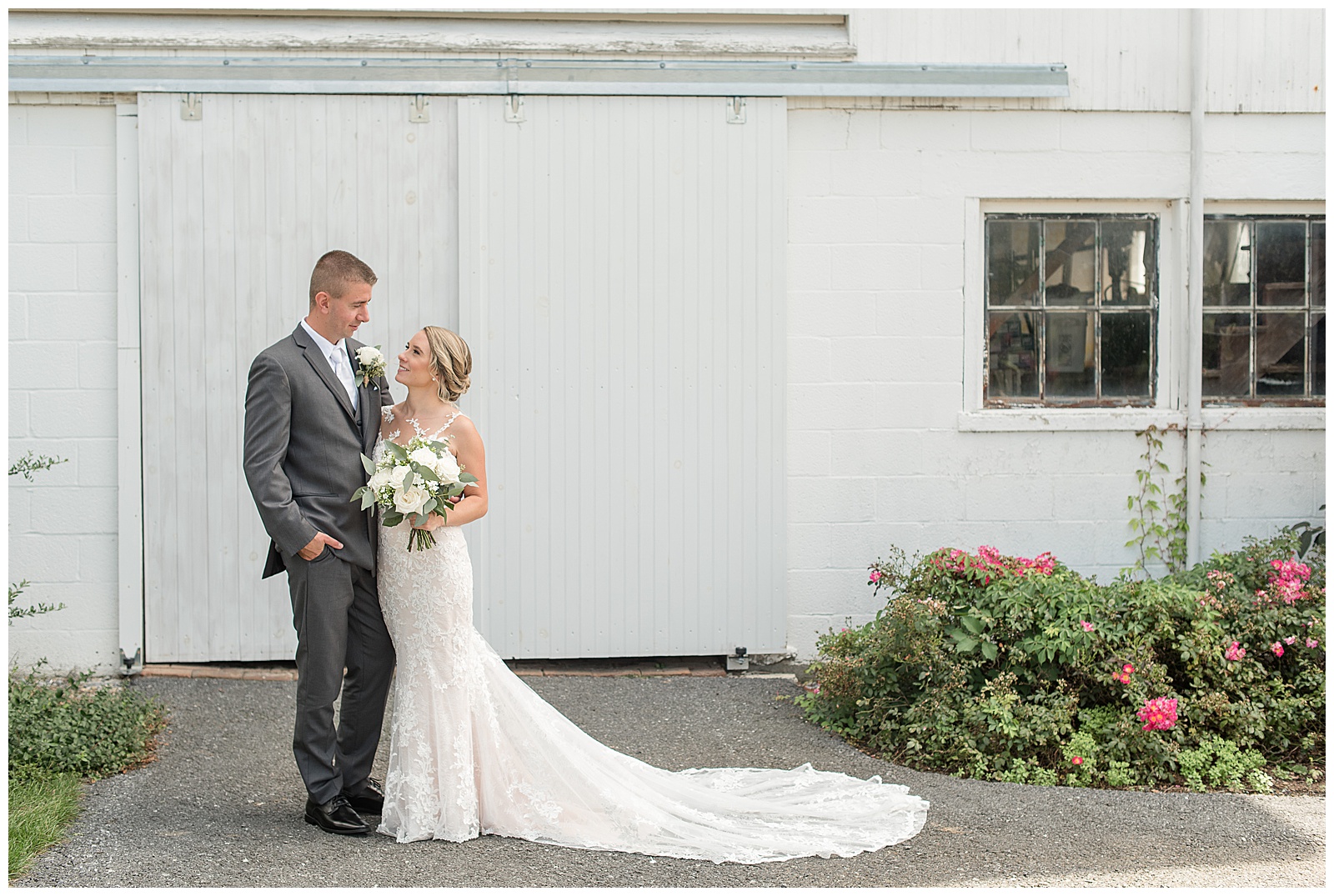the couple are on the left side of the photo standing in the driveway in front of the white barn with a large potted plant sitting on the right side of the photo on the driveway and the couple is side hugging and looking at each other while smiling with the groom on the left and the bride on the right holding her bouquet of flowers and the train of her dress is stretched out along her left side on the driveway at Lakefield Wedding in Manheim, PA