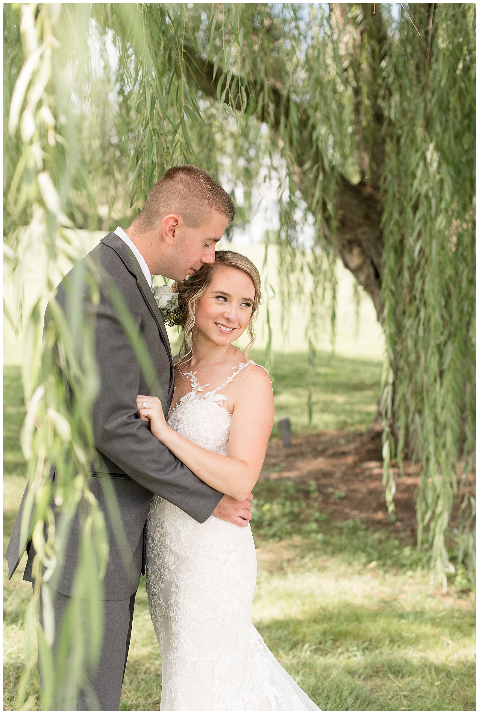 close up photo of the groom hugging the bride while he faces the right side of the photo and rests his face on the bride's side of her head and she faces him while resting her left arm on his and smiling and looking over her left shoulder while standing in front of a willow tree in a grassy field at Lakefield Weddings in Manheim, PA