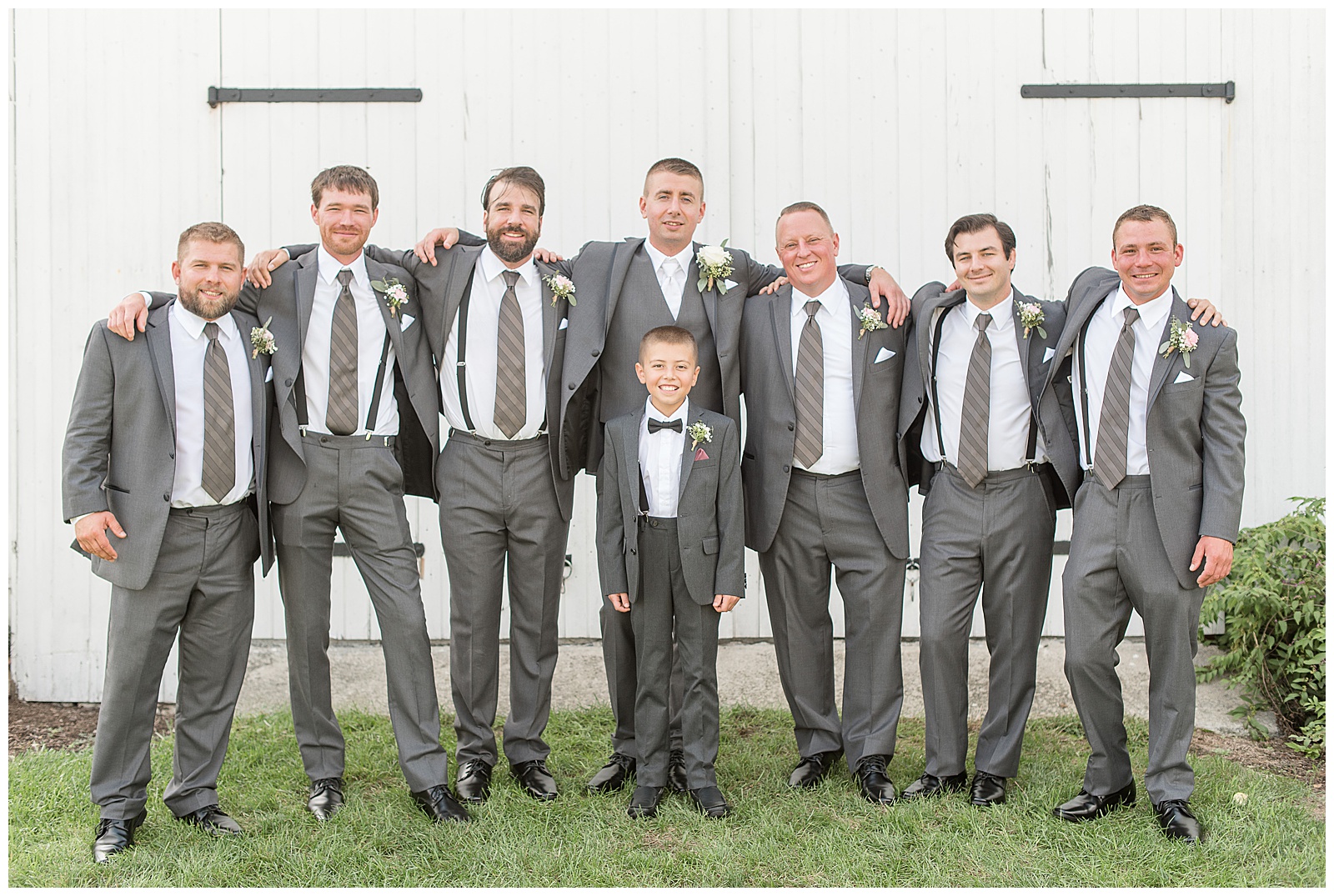 the groom is in the center with three groomsmen on his right side and three groomsmen on his left side with everyone placing their arms around the shoulders of the person next to them and the ring bearer is standing in front of the groom with his hands at his sides with everyone wearing their dark gray suits with white dress shirts and ties in front of the white barn standing in the grass at Lakefield Weddings in Manheim, PA