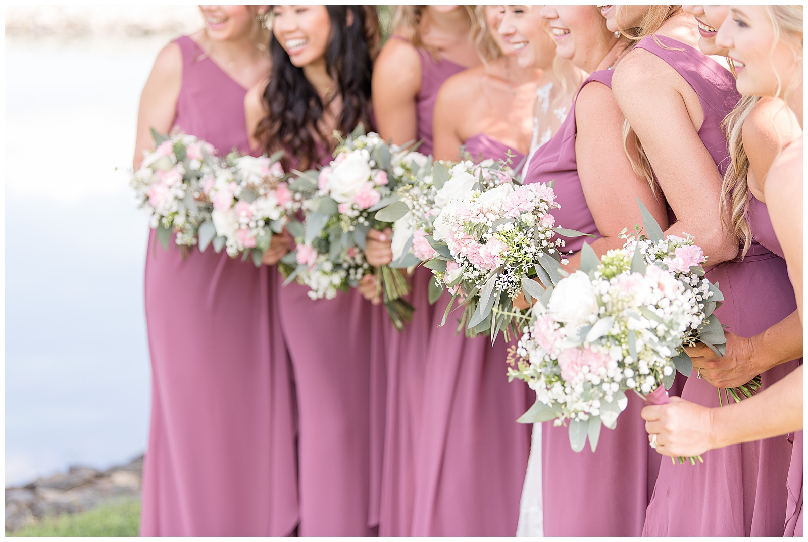 close up photo of the bridesmaids in their mauve floor-length gowns all facing towards the center and smiling and all looking off towards the left side of the photo with some of their backs slightly facing the camera and holding their beautiful pastel pink and white and green bouquets of flowers while outside on a sunny day at Lakefield Weddings in Manheim, PA