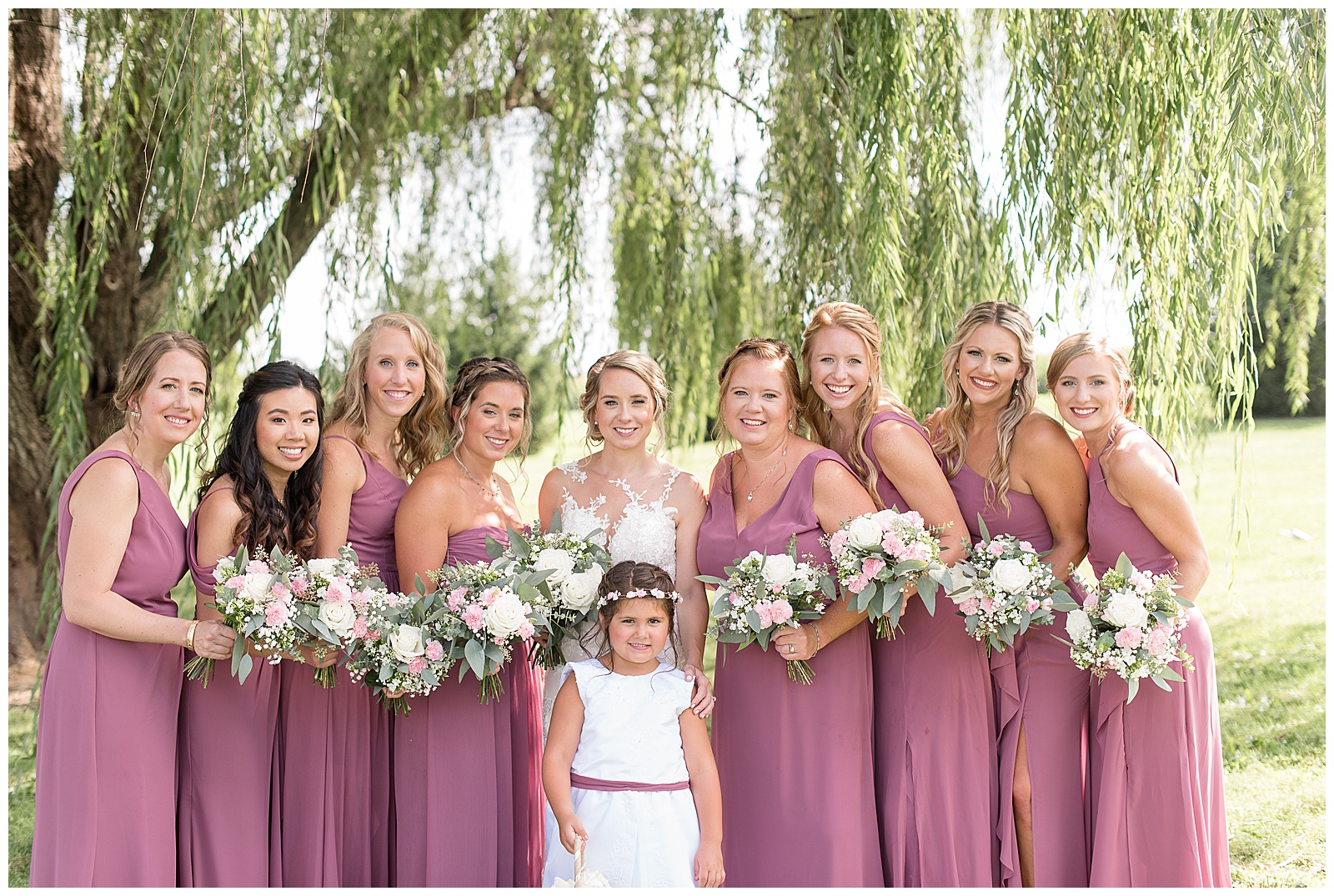 close up photo with the bride is in the center with the flower girl in front of her and four bridesmaids to the right of the bride and four bridesmaids to the left of the bride all facing towards her slightly and smiling at the camera while holding their flower bouquets and standing under the willow tree on a sunny day at Lakefield Weddings in Manheim, PA