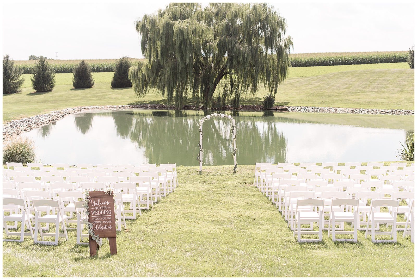 a view of the ceremony location with no guests in the picture with rows of white chairs on each side of the aisle and a small wooden welcome side to the left of the aisle and the chairs face a beautiful pond with willow tree-lined grassy field with cornfield behind that on a sunny day at Lakefield Weddings in Manheim, PA
