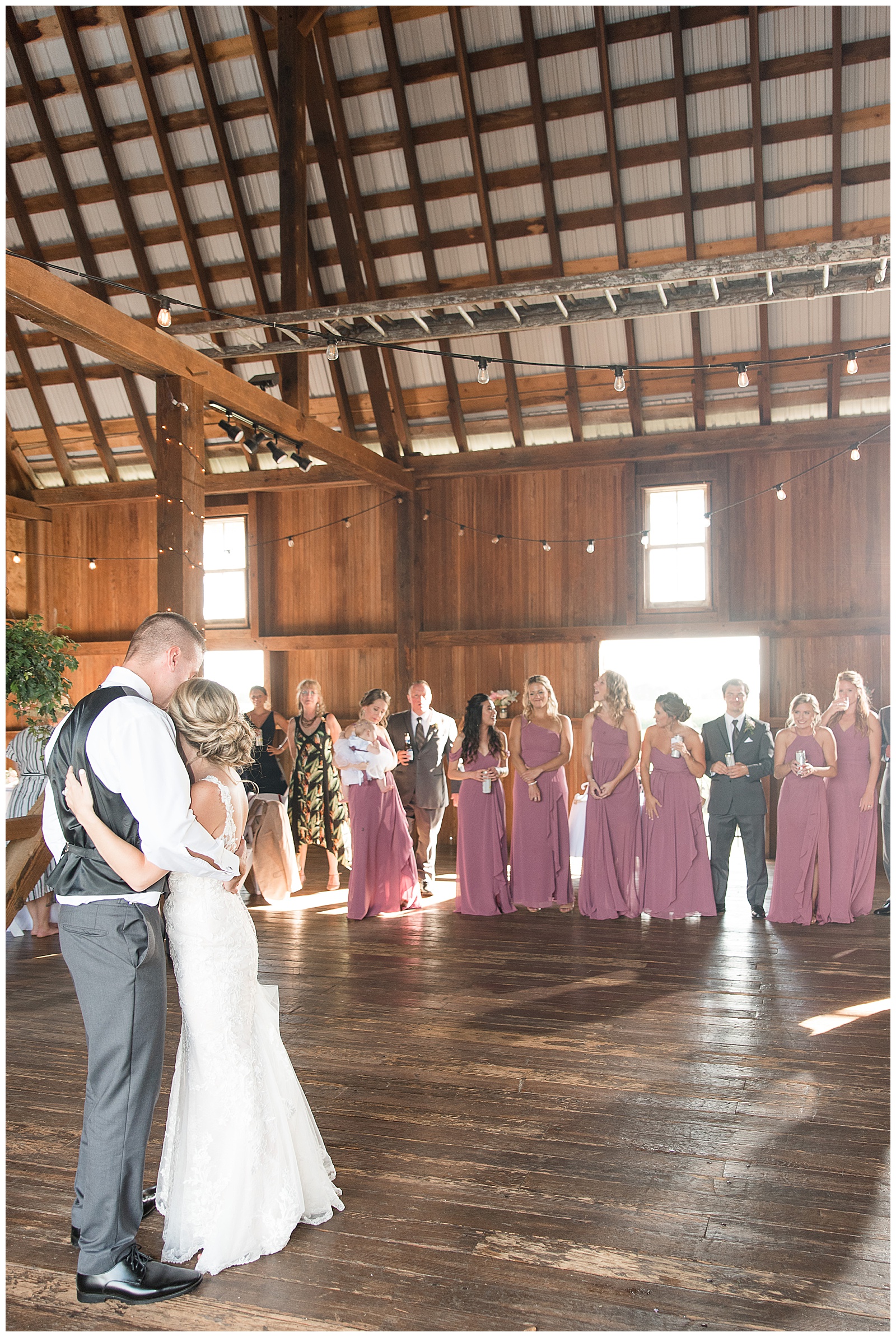 the groom is on the left and the bride on the right and they are hugging one another and slow dancing with their backs to the camera on the left side of the photo with their bridal party lined up further behind them watching in the barn reception area at Lakefield Wedding in Manheim, PA