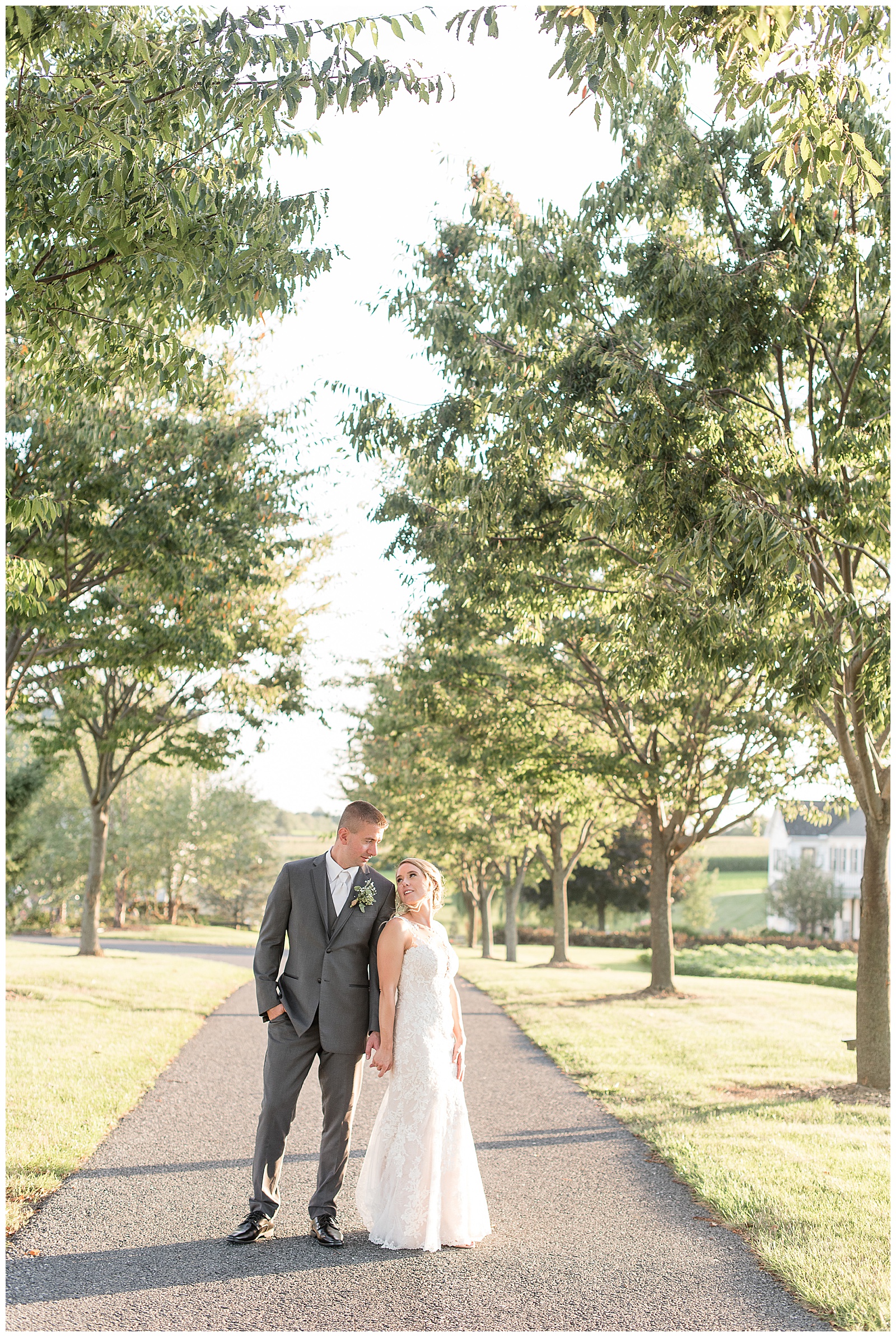 the couple is standing on a paved tree-lined pathway outdoors on a sunny day with the groom on the left with his right hand in his pocket and leaning towards the bride on the right whose back is slightly towards him while she looks up at him and they both are smiling at Lakefield Weddings in Manheim, PA