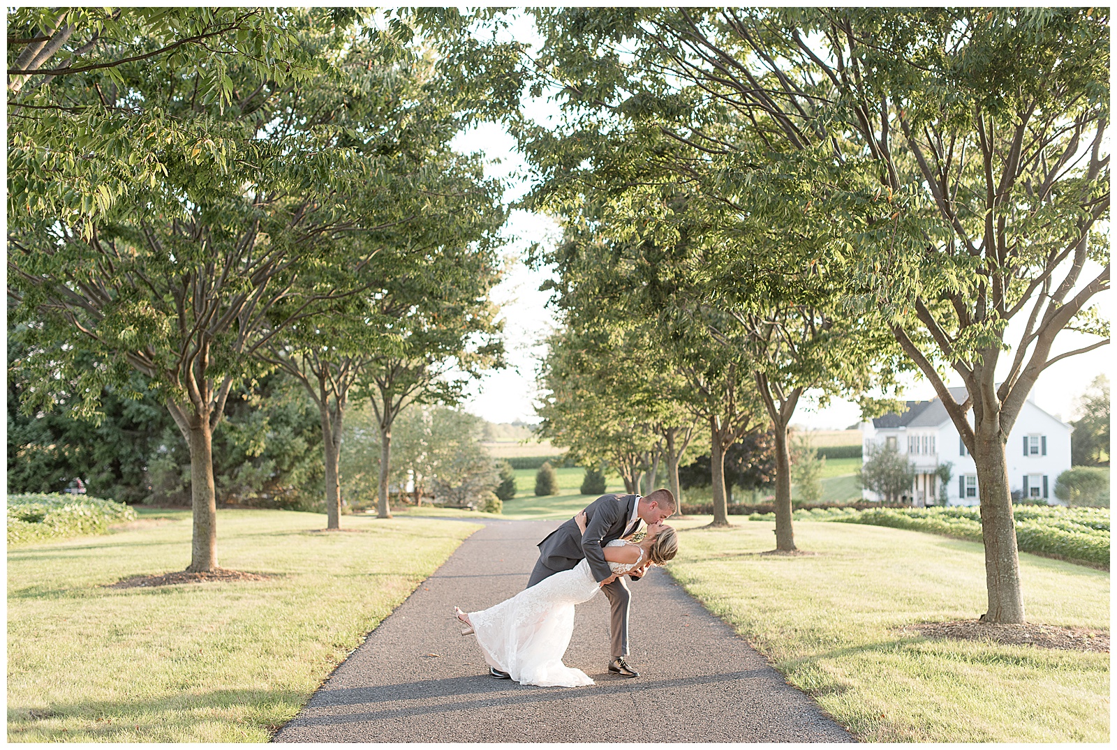 the couple is standing in the center of a tree-lined paved path with the groom standing behind the bride and facing right while dipping her backwards as they kiss on a sunny day at Lakefield Weddings in Manheim, PA