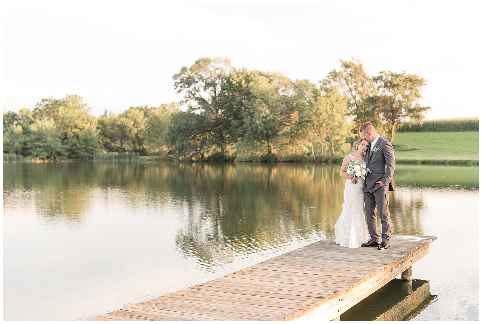 the couple is standing at the end of the dock on the pond with trees behind them and the bride is on the left holding her bouquet and resting her head on the groom and the groom is standing on the right with his left hand in his pocket on a sunny day at Lakefield Weddings in Manheim, PA