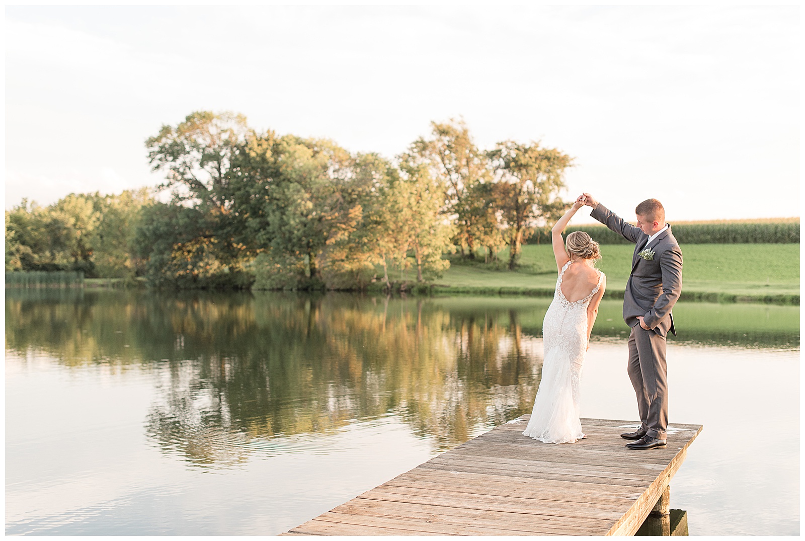 groom is twirling the bride under his right arm while holding her left hand at the end of the dock at the pond with the trees reflecting in the pond behind them on a sunny day at Lakefield Weddings in Manheim, PA