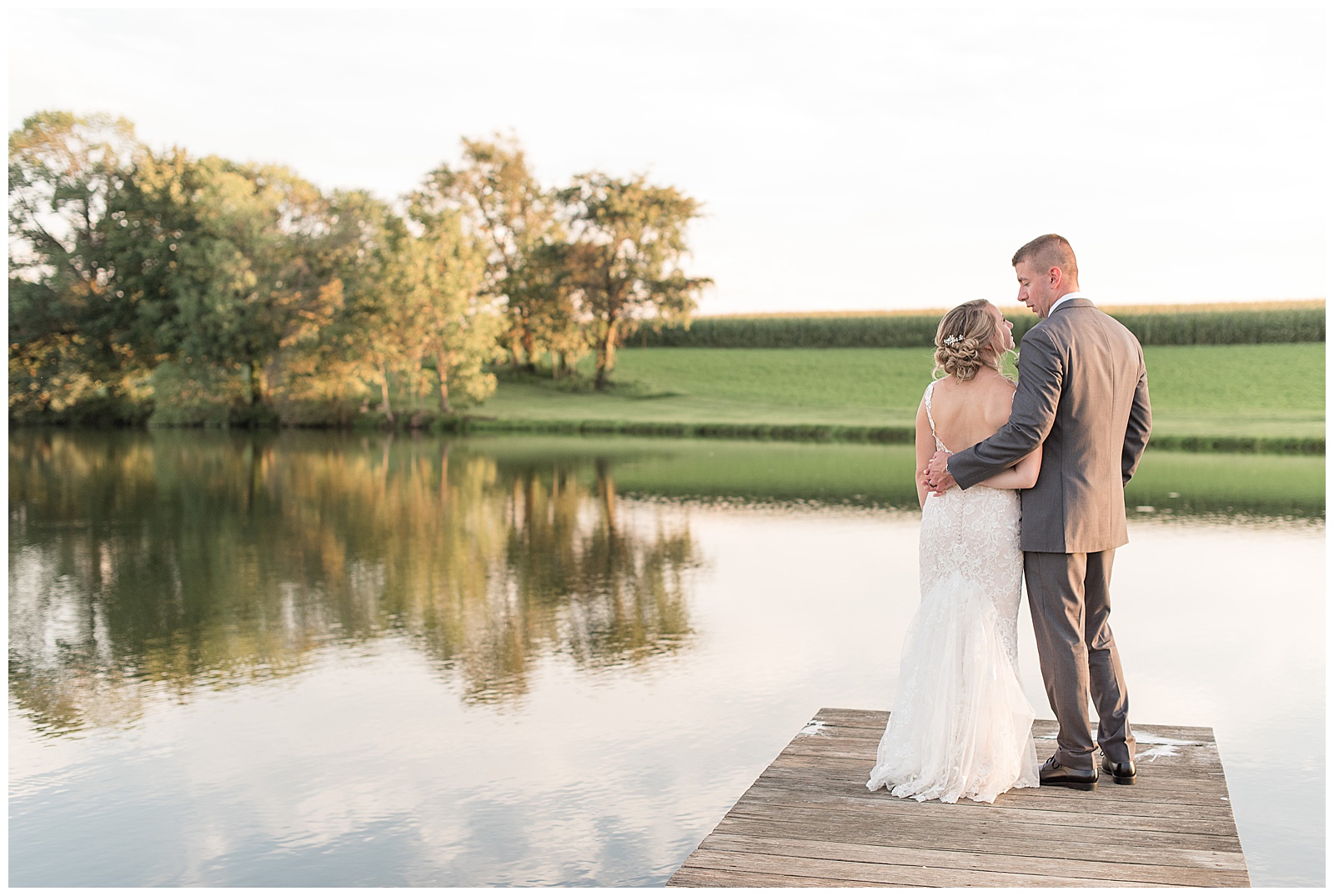 the couple is standing at the end of the dock with the bride on the left and the groom on the right with their backs to the camera and arms around one another looking at each other with the pond and tree reflections in the background on a sunny day at Lakefield Weddings in Manheim, PA