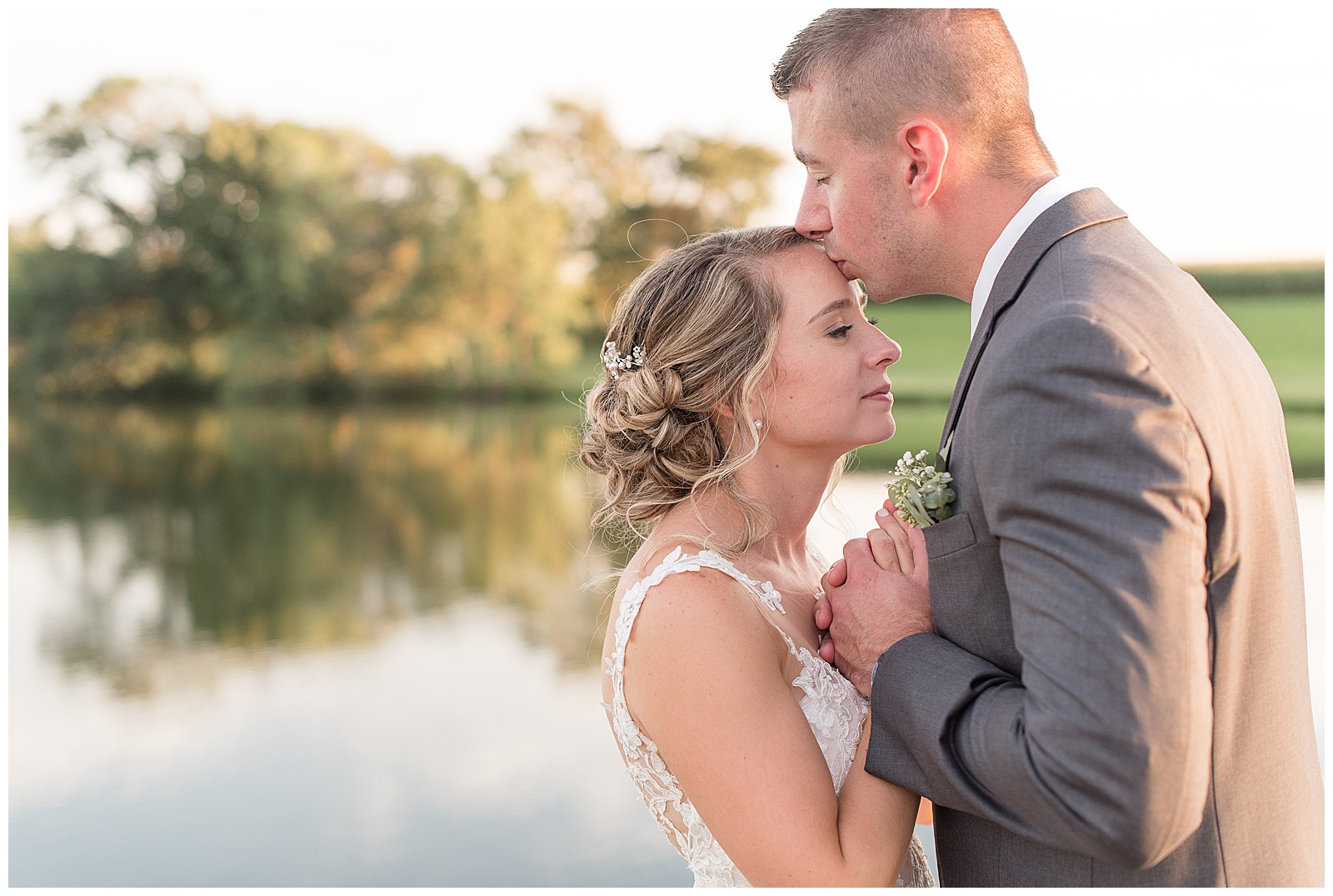 close up of the groom kissing the bride's forehead as she smiles and closes her eyes and she is on the left and the groom is on the right with the pond and trees behind them at Lakefield Weddings in Manheim, PA
