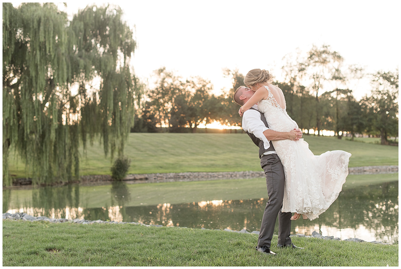 the couple is on the right side of the photo with the pond and trees and sunset behind them as the groom is on the left and is lifting the bride off the ground while they kiss each other at Lakefield Weddings in Manheim, PA