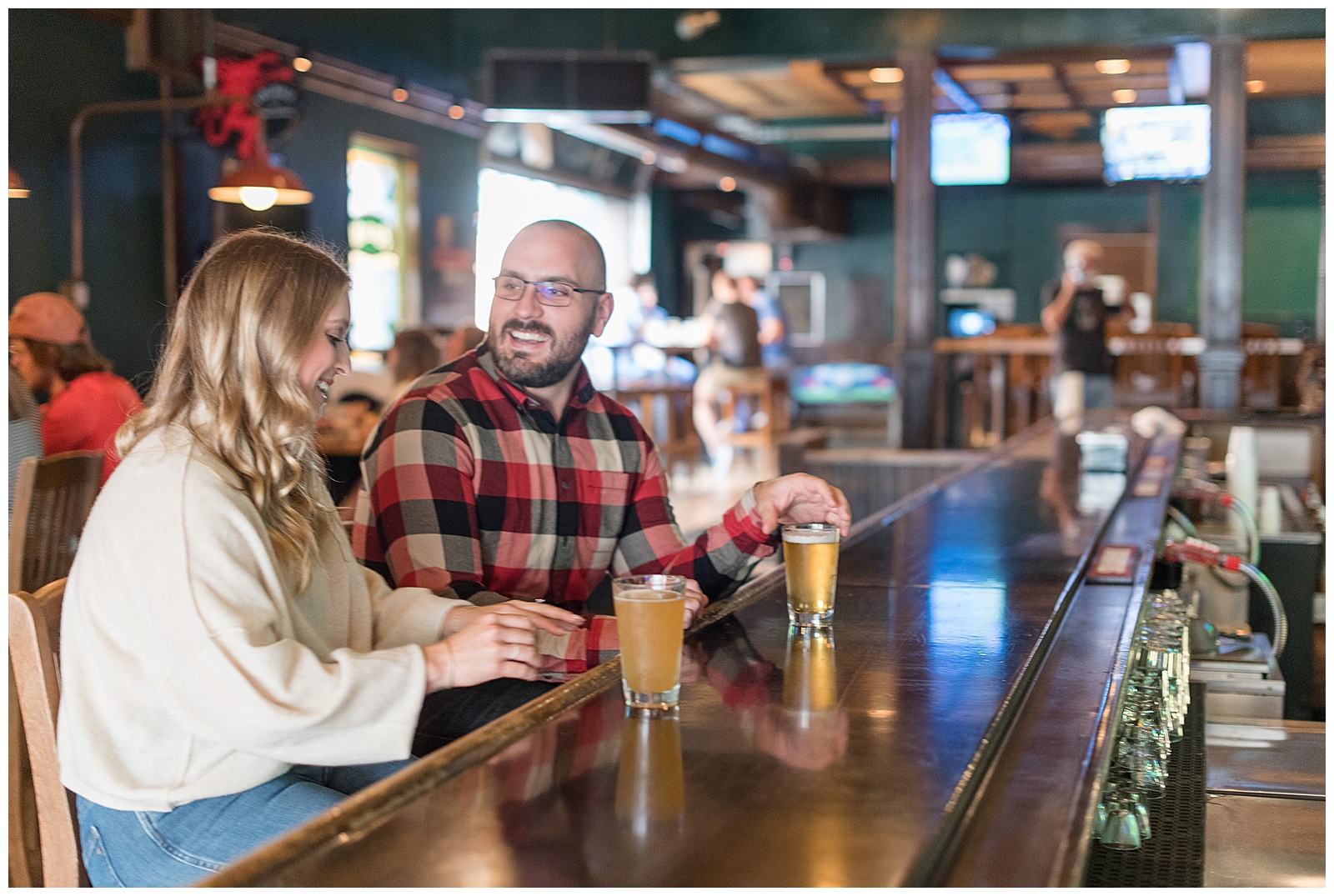 couple is sitting at the bar with drinks on the counter in front of them and the girl is on the left looking down and reaching for her drink smiling and the guy is on the right turning towards the girl smiling and reaching for his drink with bright light shining in the doorway behind them in downtown Pittsburgh