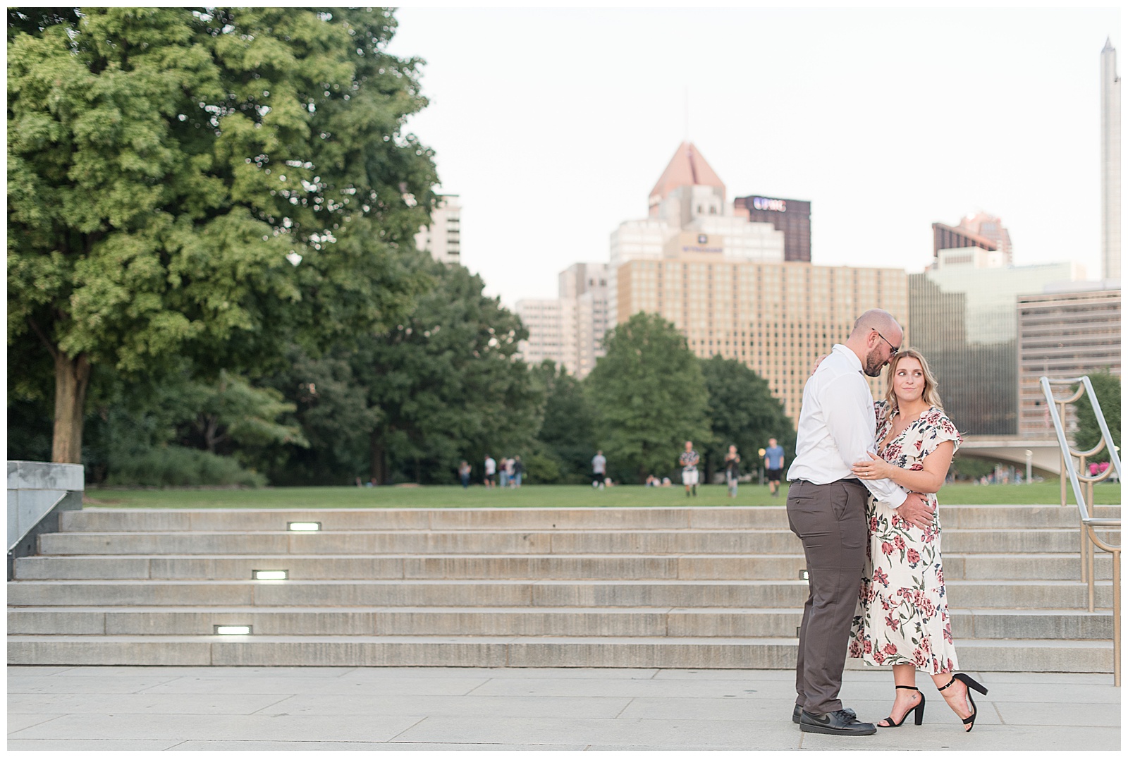 the couple is on the right hand side of the photo facing each other at the bottom of a small set of concrete steps outdoors with large trees and tall buildings behind them and the guy has his arms around the girl and his resting his forehead on the top of the girl's head as she is looking over her left shoulder with her arms also around him in downtown Pittsburgh