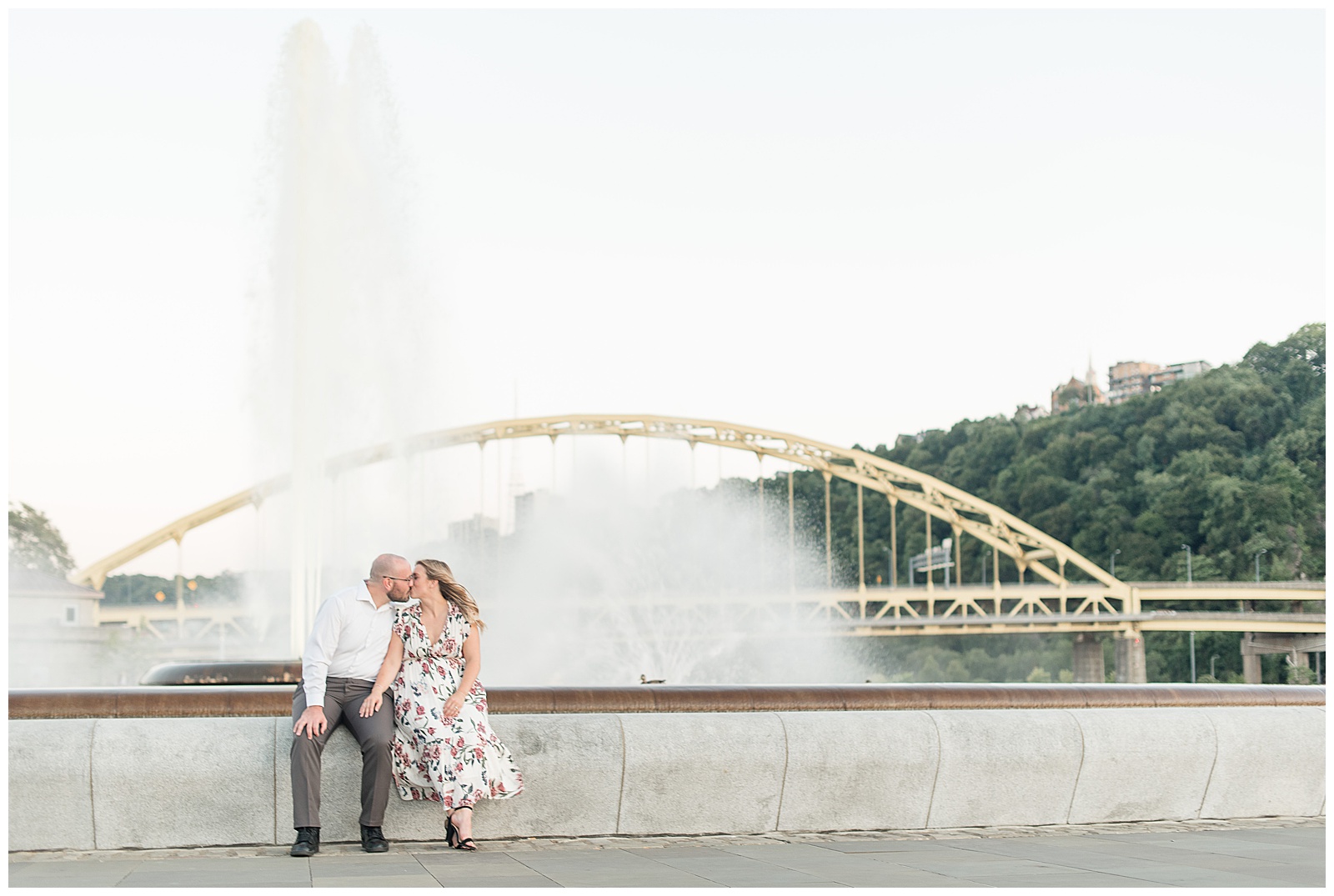 the couple is outdoors sitting along a concrete wall in front of a water fountain and white steel bridge with trees behind it and the guy is on the left and the girl is on the right and they are sitting close together and kissing as the girl rests her right hand on the guy's left knee in downtown Pittsburgh