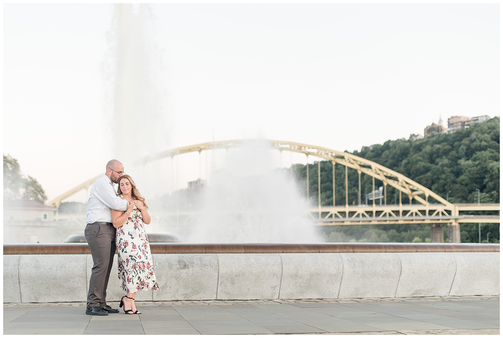 the couple is on the left hand side of the photo with the concrete wall and water fountain and white steel bridge and trees behind them and the guy is on the left turning towards the girl and embracing her with his arms around her shoulders as she faces the camera and leans into him and smiles in downtown Pittsburgh