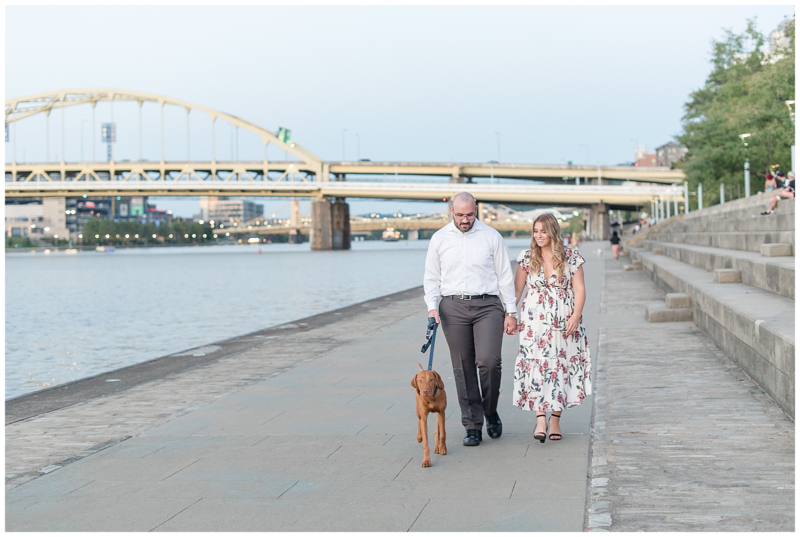 the couple is walking along the river on the concrete walkway towards the camera with the bridge and river behind them as the guy is on the left holding the dog's leash and the dog is walking ahead of the couple and the girl is on the right and the couple is holding hands and looking down towards the dog in downtown Pittsburgh