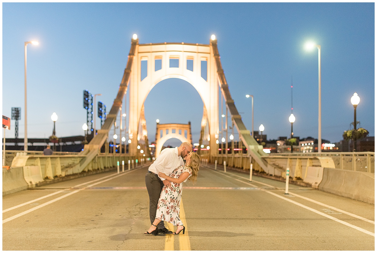 the couple is standing in the middle of the road at dusk with a lit-up arching bridge and streetlights behind them and the guy is on the left dipping the girl backwards and holding her as they kiss and her left foot reaches out to point towards the ground in downtown Pittsburgh