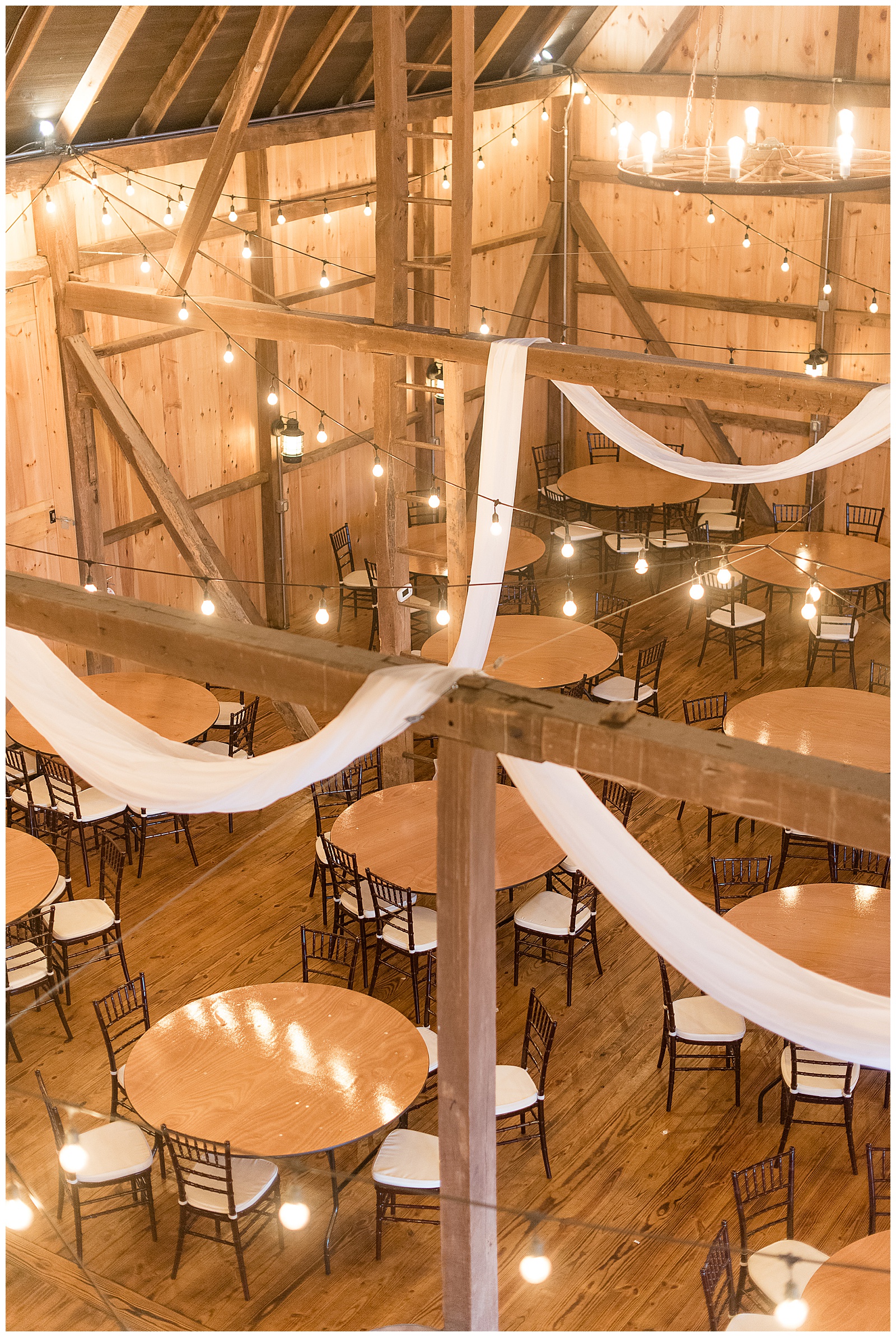 close up interior photo of rustic barn venue with angle of photo from higher up inside the barn looking down over the wooden rafters and stringed lights and draped fabric above the tables and chairs at Spring Valley Farms in Dover, PA