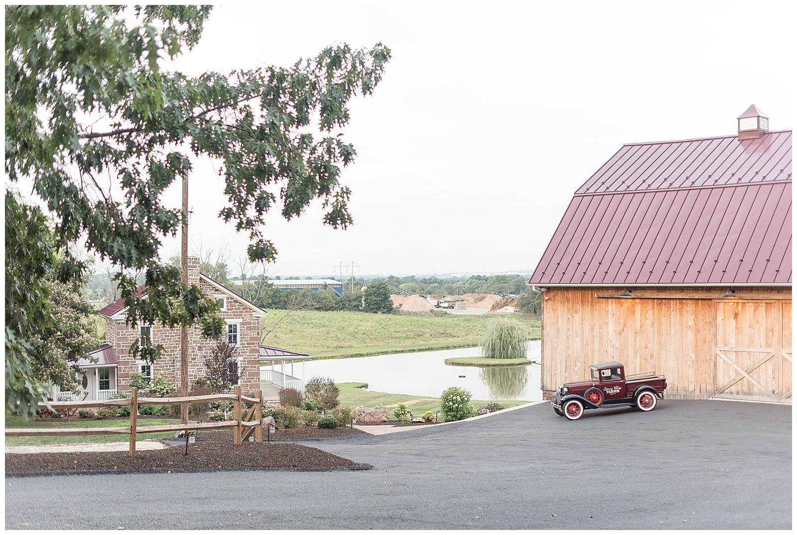 photo on driveway with views of tree branches and sandstone farmhouse to the left and the pond and valley and grass yard in the center and the maroon Model A Ford parked in front of the rustic large barn with maroon metal roof to the right of the photo at Spring Valley Farms in Dover, PA